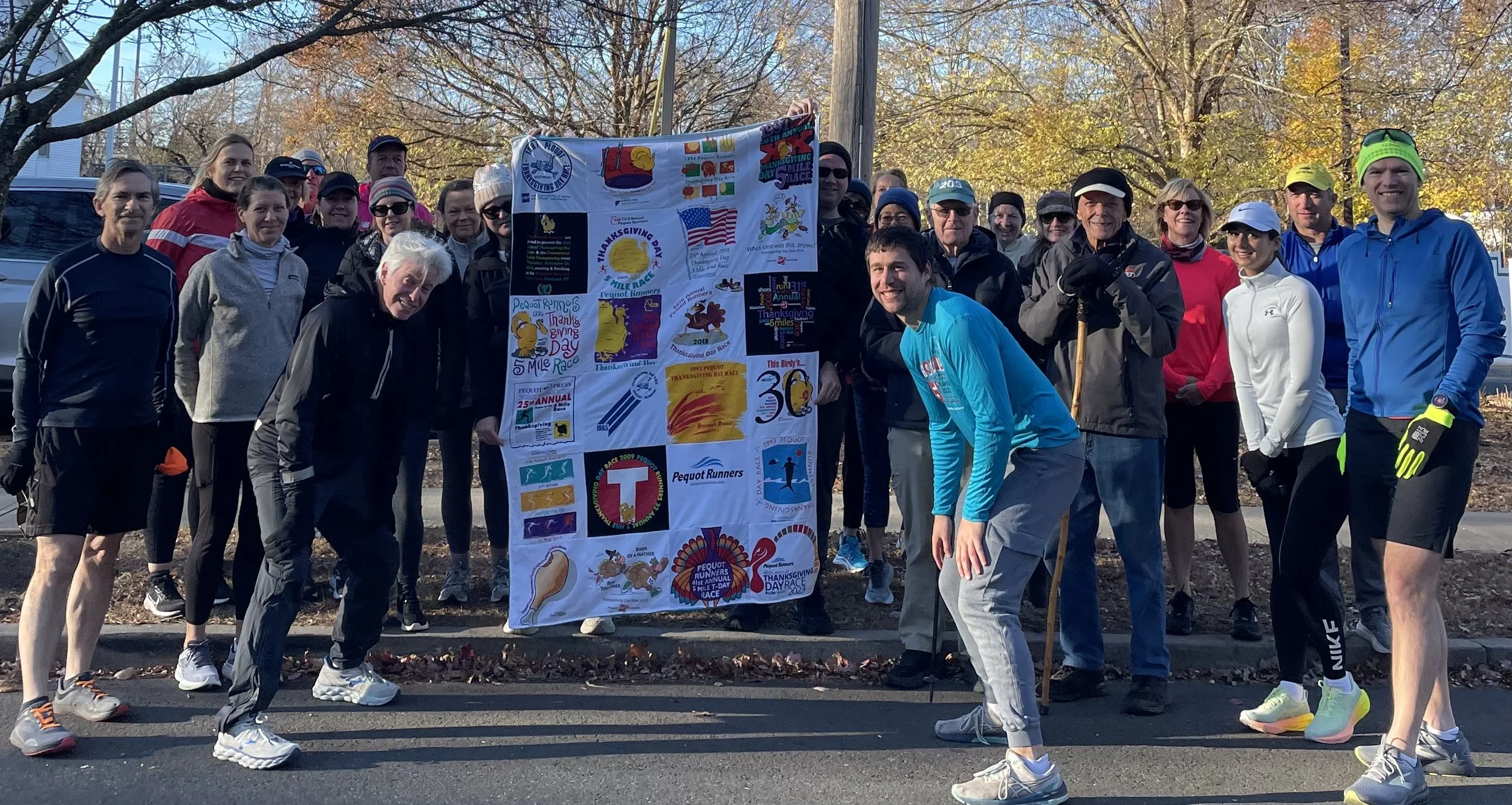 Group of people participating in a Thanksgiving Day race, holding a commemorative banner, outdoors on a fall day with trees in the background.
