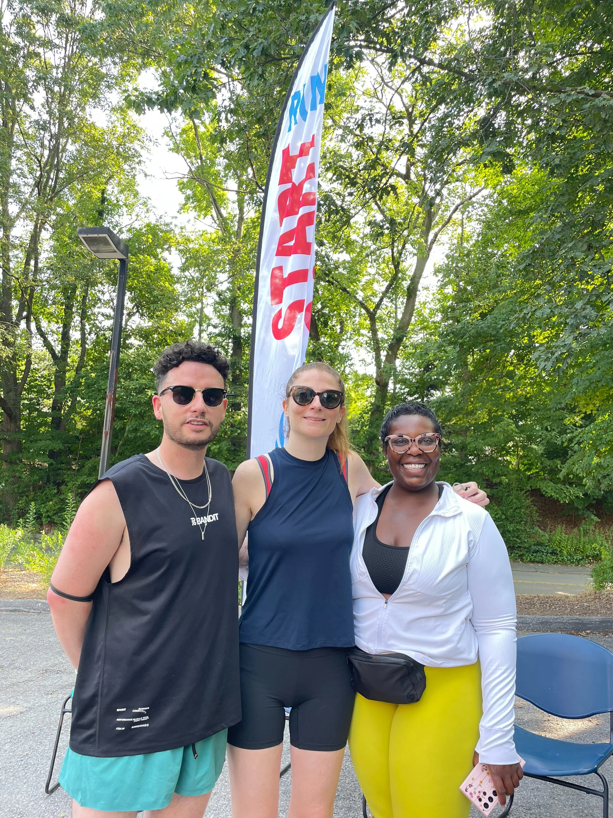 Three people standing outdoors with a flag that reads "START" in the background, surrounded by green trees, smiling at the camera.