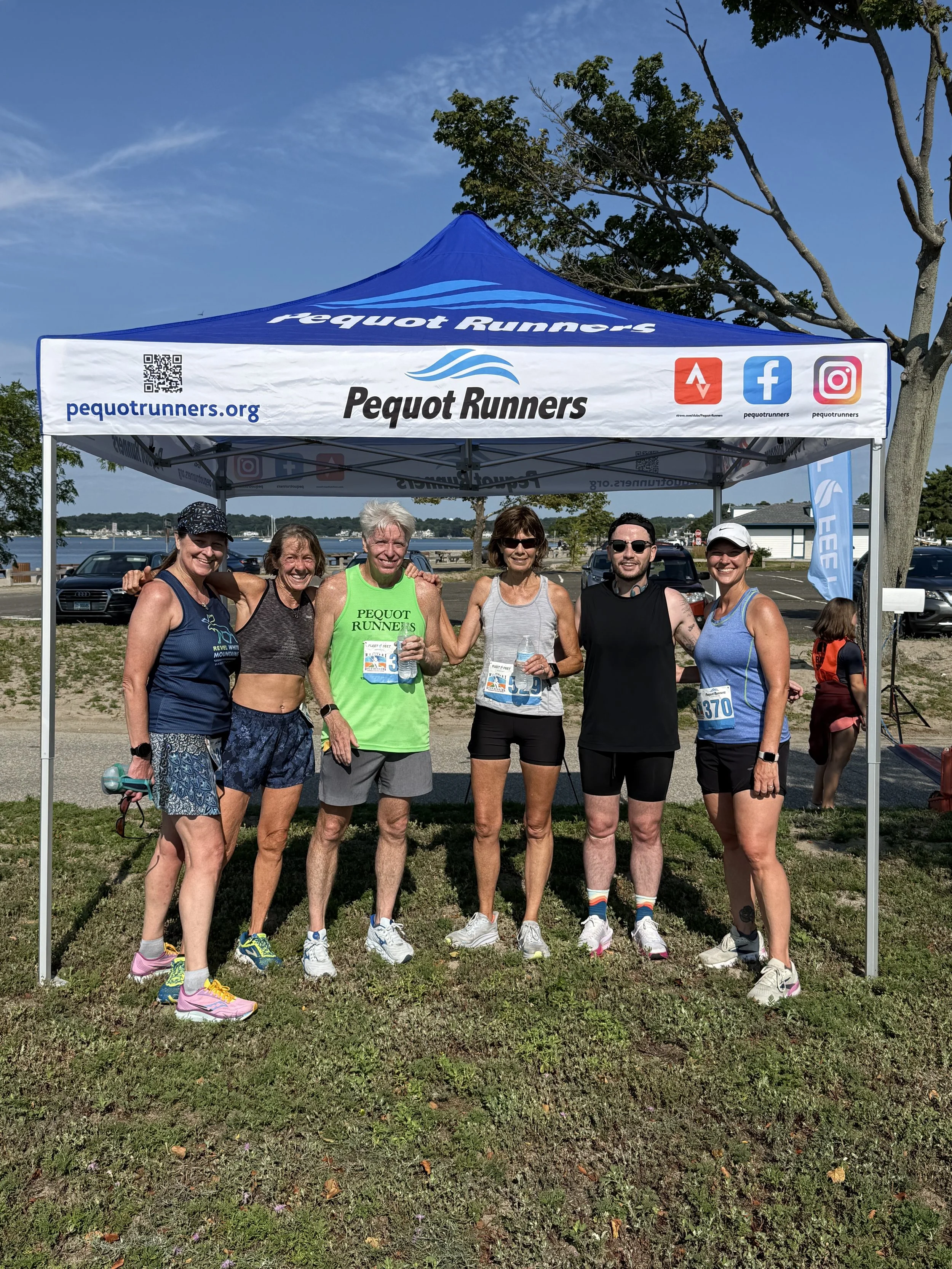 Group of six runners standing together under a blue tent at a park, smiling after a race, with a body of water and trees in the background.