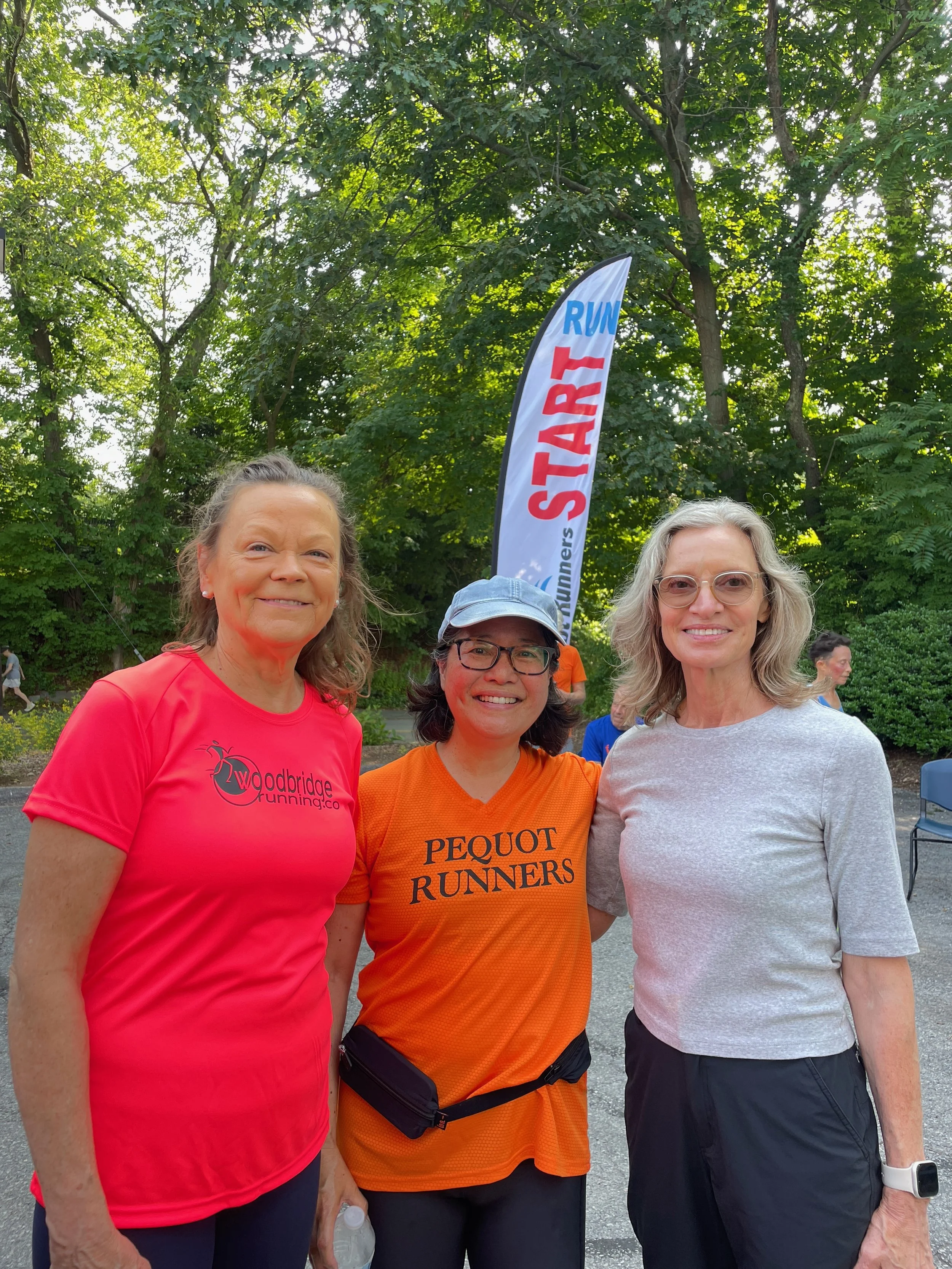 Three women standing outdoors in front of a 'START' running banner, smiling at the camera, with trees in the background.