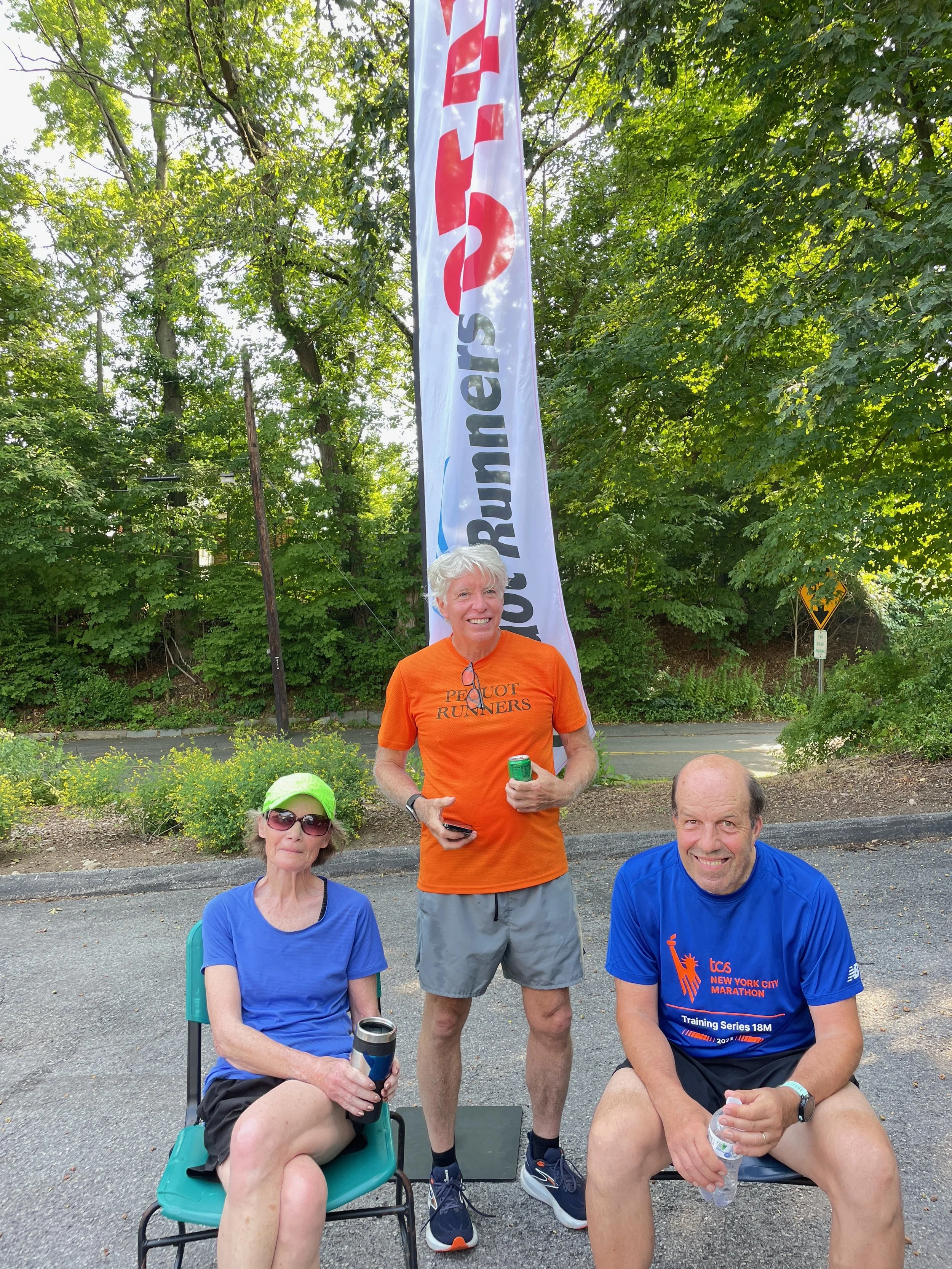 Three people, one woman and two men, are outdoors in a park-like setting with trees and greenery. The woman is seated on a chair, wearing sunglasses, a blue shirt, and a bright green cap, holding a water bottle. The man on the right is seated on a ch