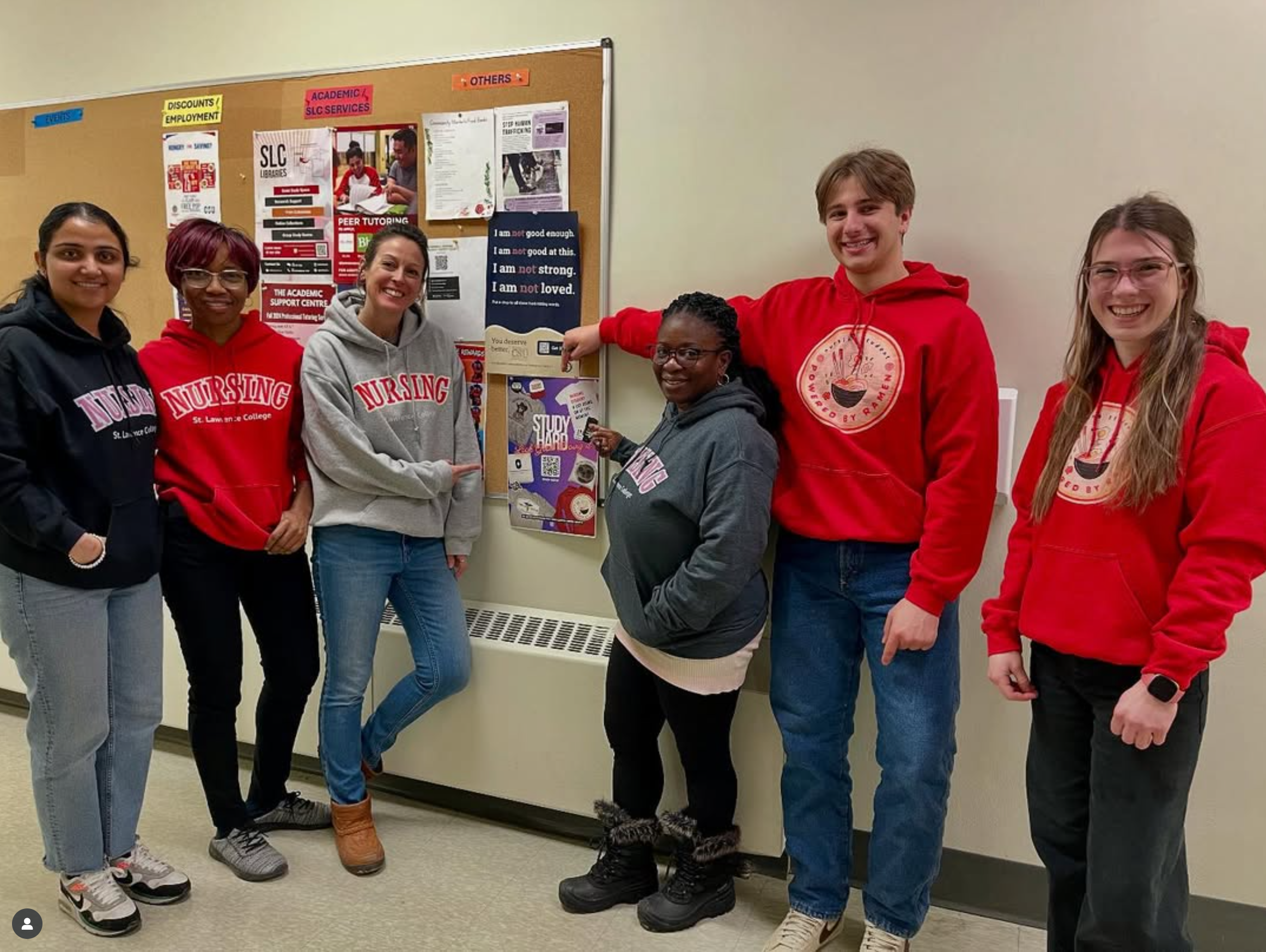 Group of six students standing in front of a bulletin board, pointing at the Artful Nurse Poster, some wearing red hoodie or sweatshirt with nursing logos, smiling at the camera.