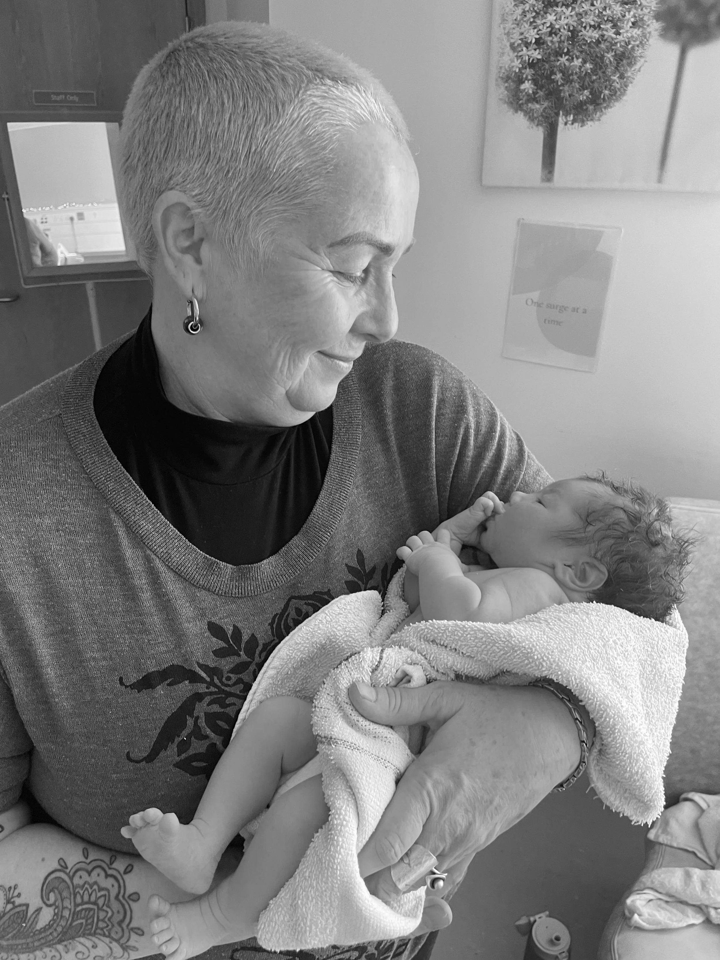 An elderly woman with short hair and earrings holding a baby wrapped in a towel and blanket in her arms indoors.