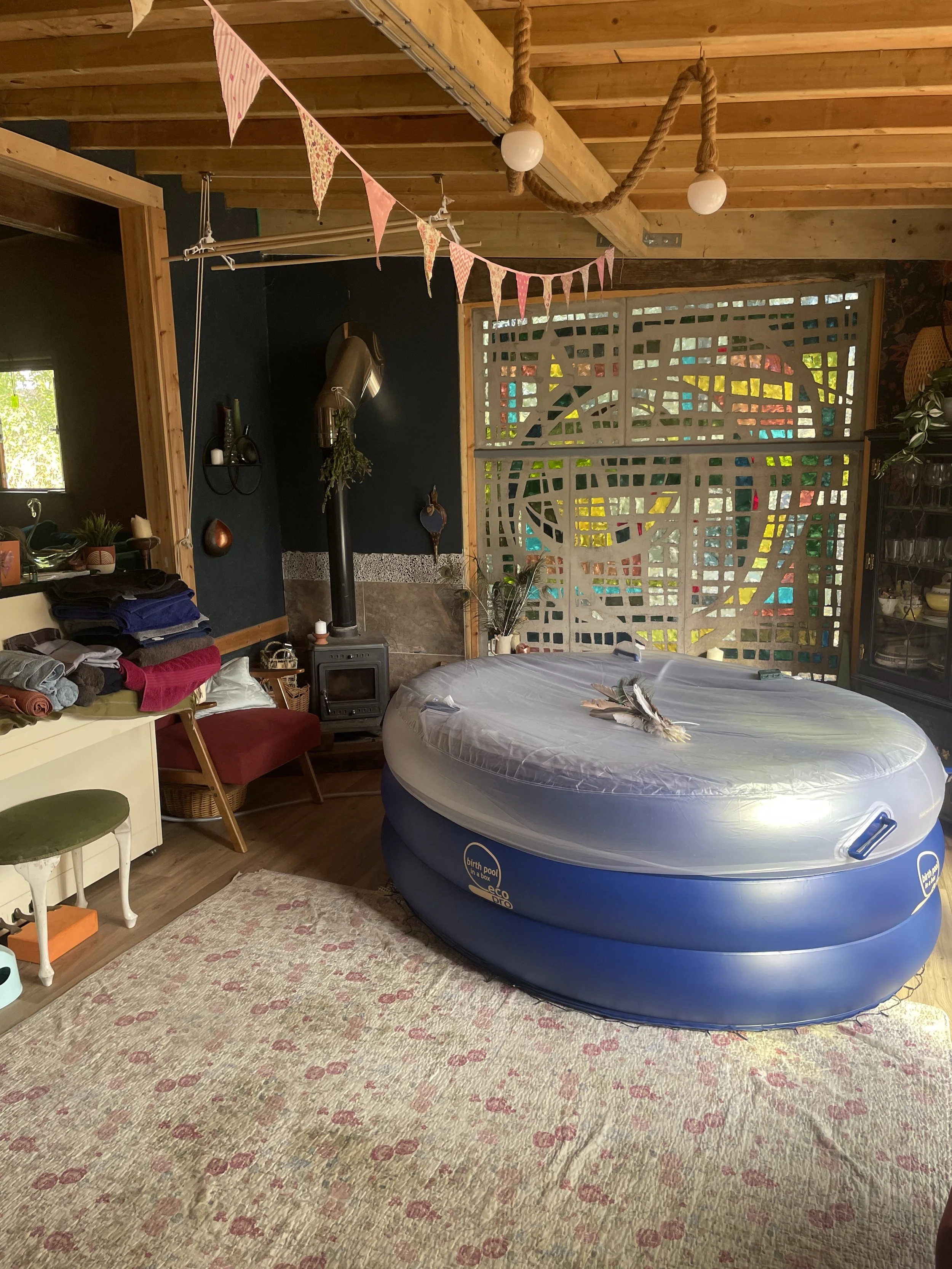 Interior of a cozy room with a large blue inflatable pool on a floral area rug, surrounded by a variety of furniture and decorations. The ceiling is made of wood beams, and colorful triangular pennant banners hang across the room. A shelving unit wit