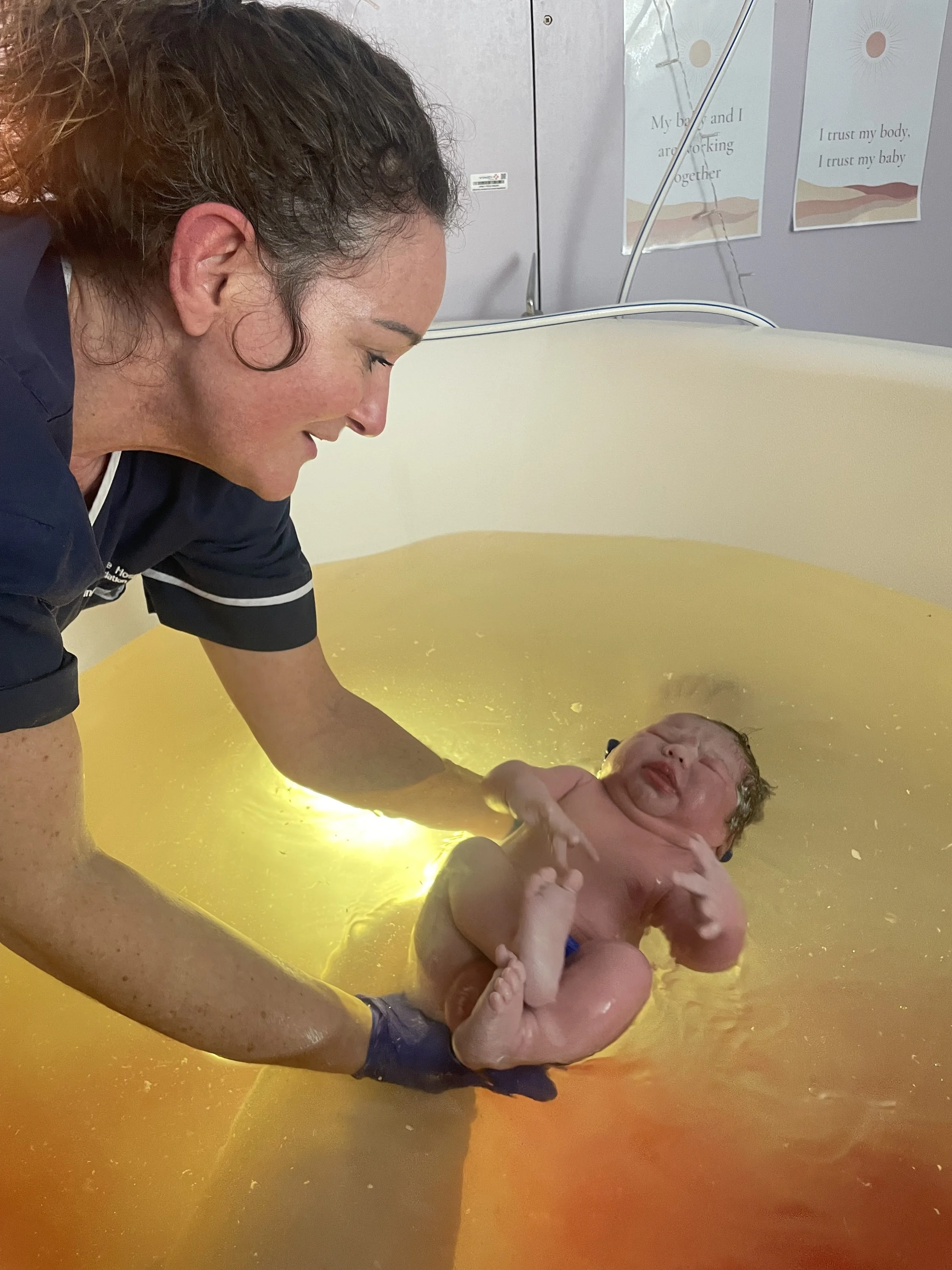 A woman assists a newborn baby during a water birth in a yellow-tinted birthing pool in a hospital setting.