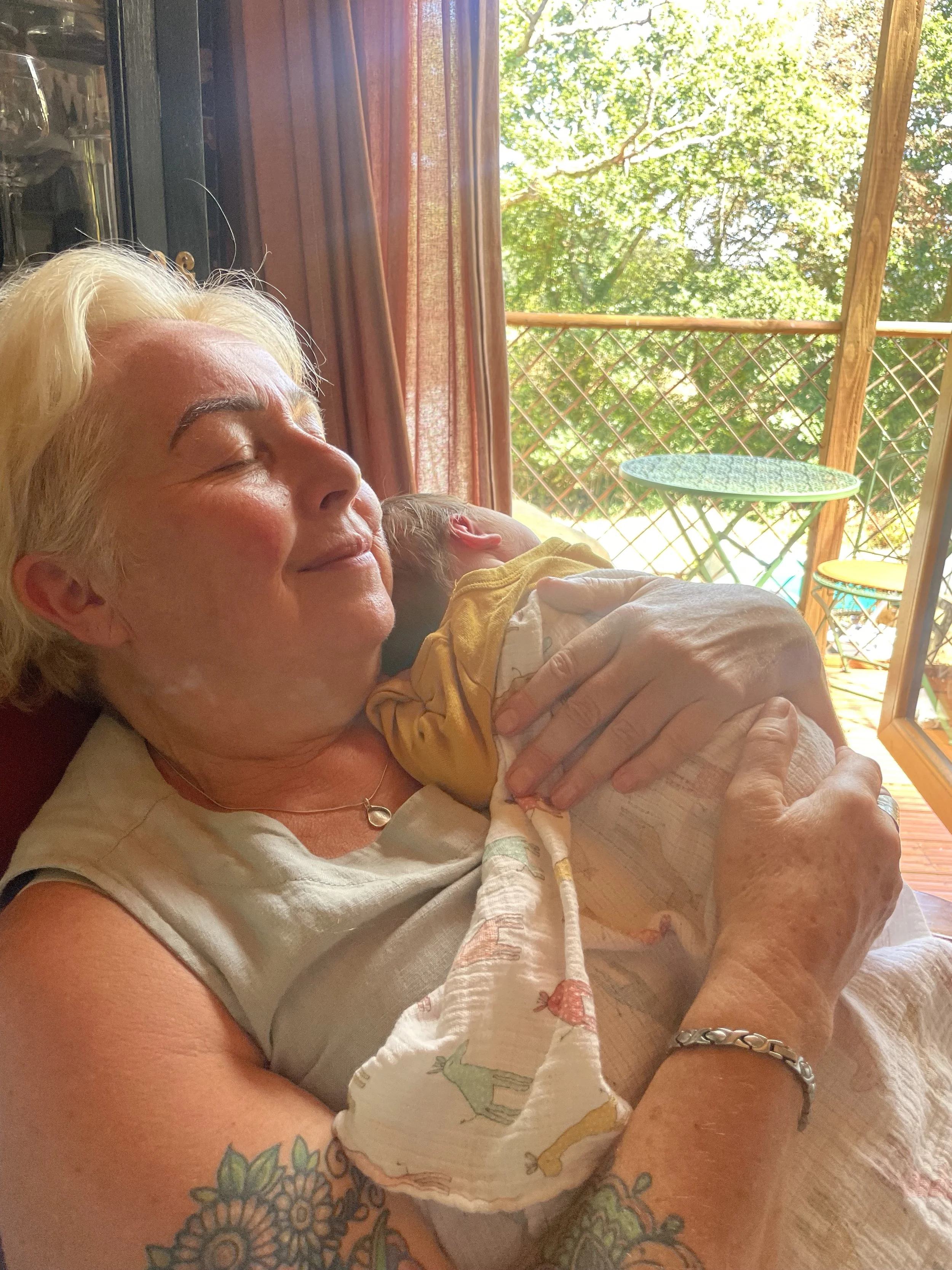 An older woman with blond hair and tattoos on her arm is lying on a couch, smiling with her eyes closed, holding a sleeping child who is resting on her chest. The background shows a balcony with a table and chairs, and trees outside.