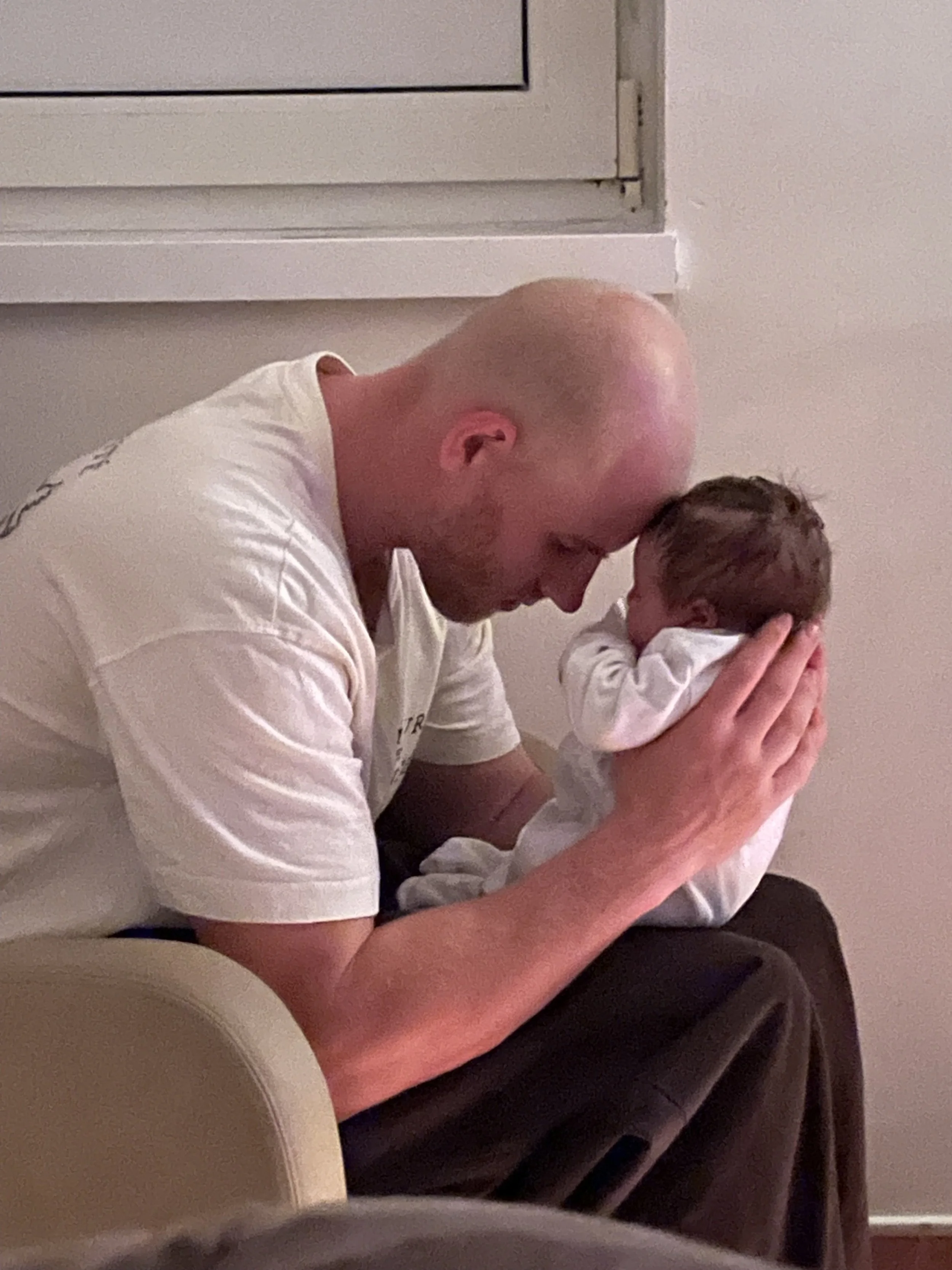 A bald man in a white shirt and a young boy with dark hair are sitting on a beige couch against a plain wall. Both are holding and looking at a smartphone.