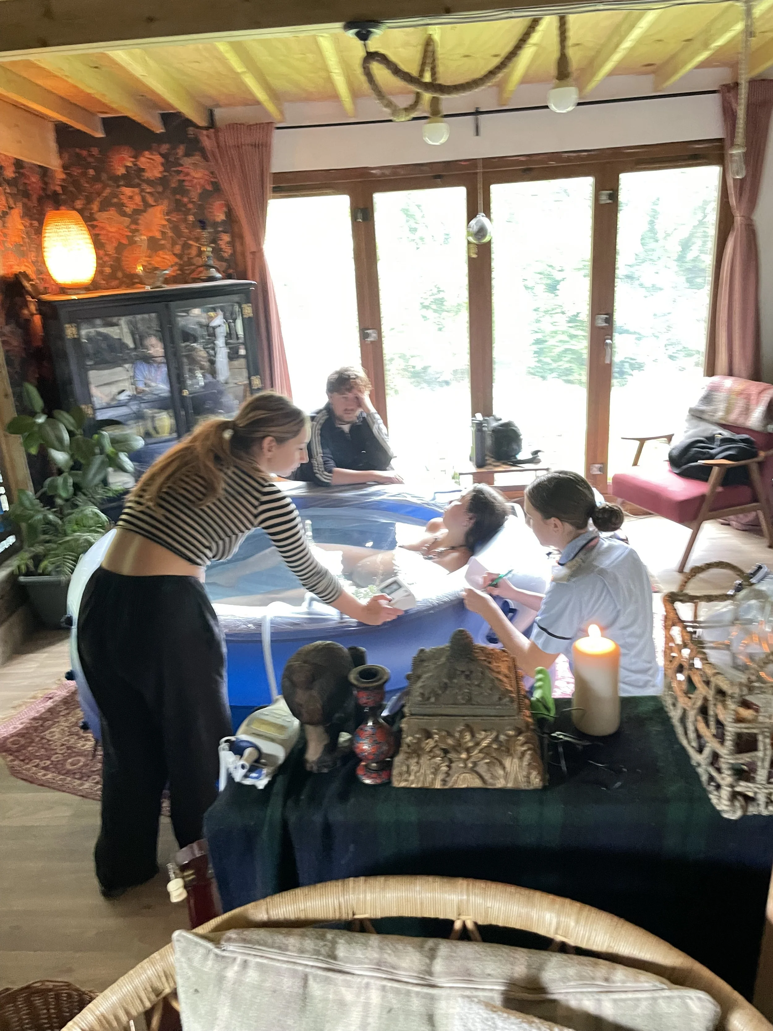 People in a cozy room gather around an inflatable pool, likely for a relaxing or recreational activity. The room features wooden walls, large windows, and various decorative items, including a lit lamp, plants, and furniture.