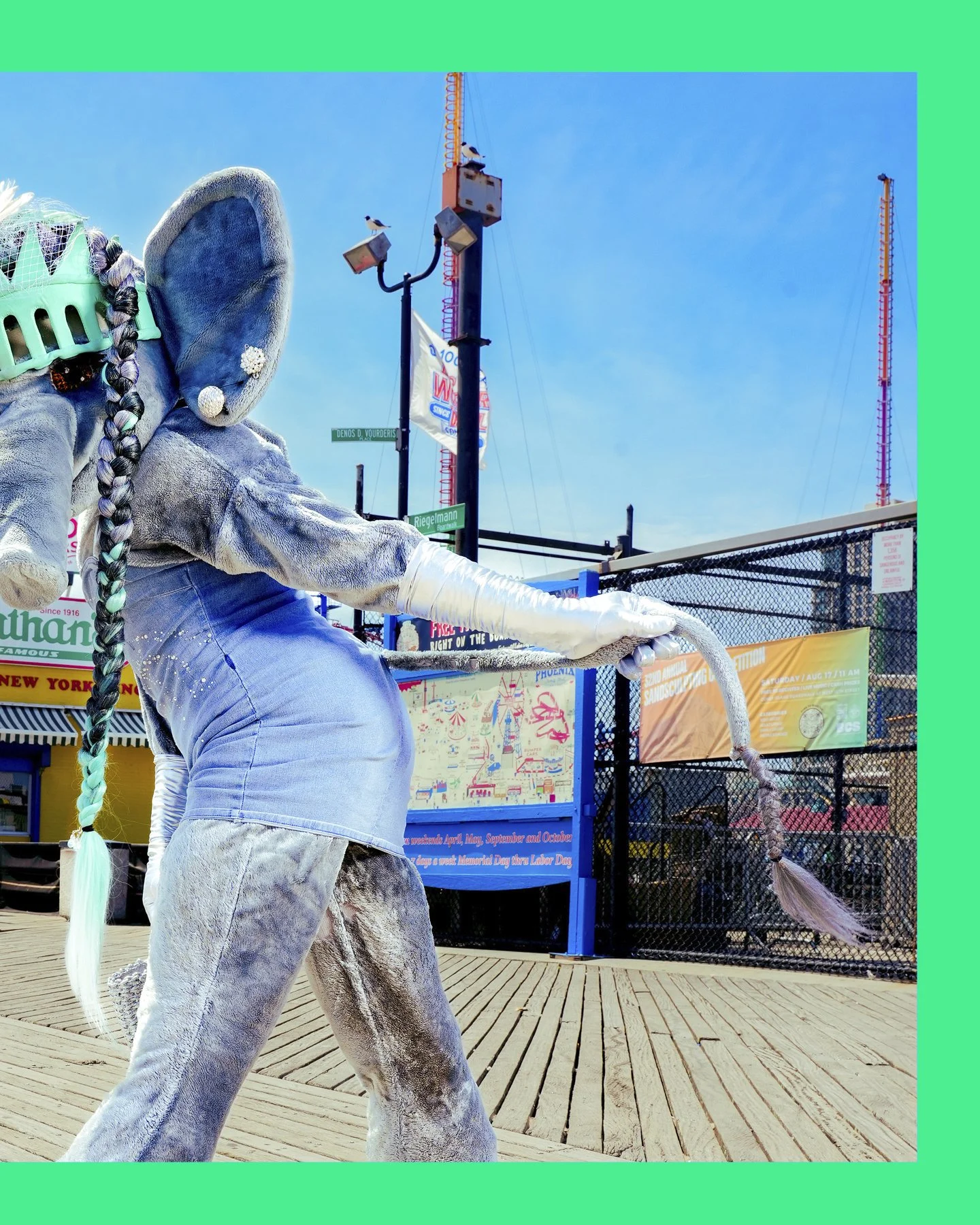 Person dressed in a grey costume with a braided hair extension, wearing a denim skirt and a green hairnet, sitting on a wooden platform near a chain-link fence at an amusement park or fairground.
