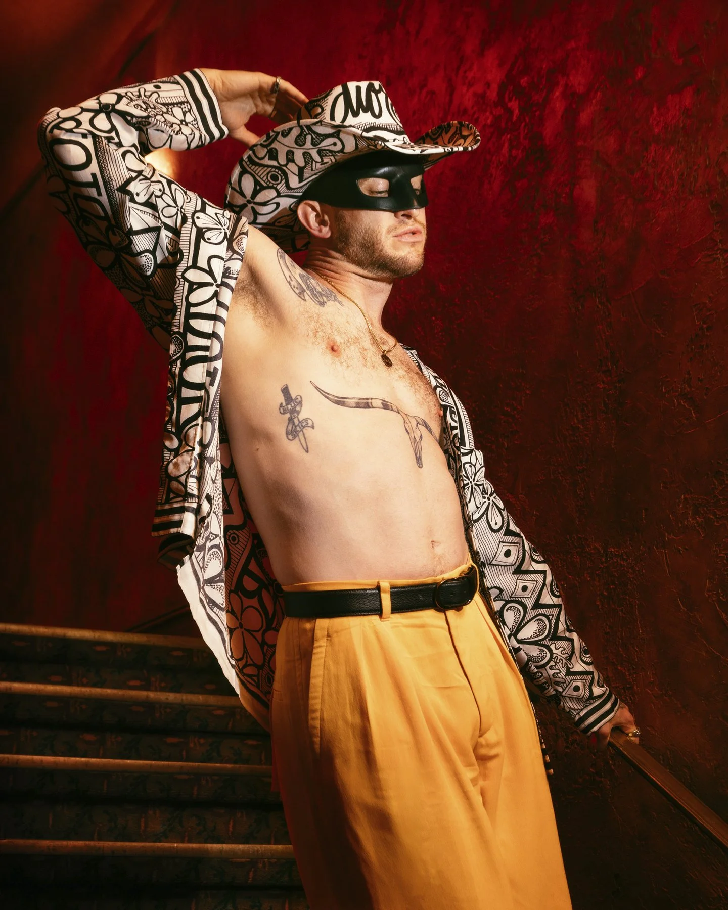 A man wearing a patterned hat and jacket, black mask, yellow pants, posing on stairs against red wall.