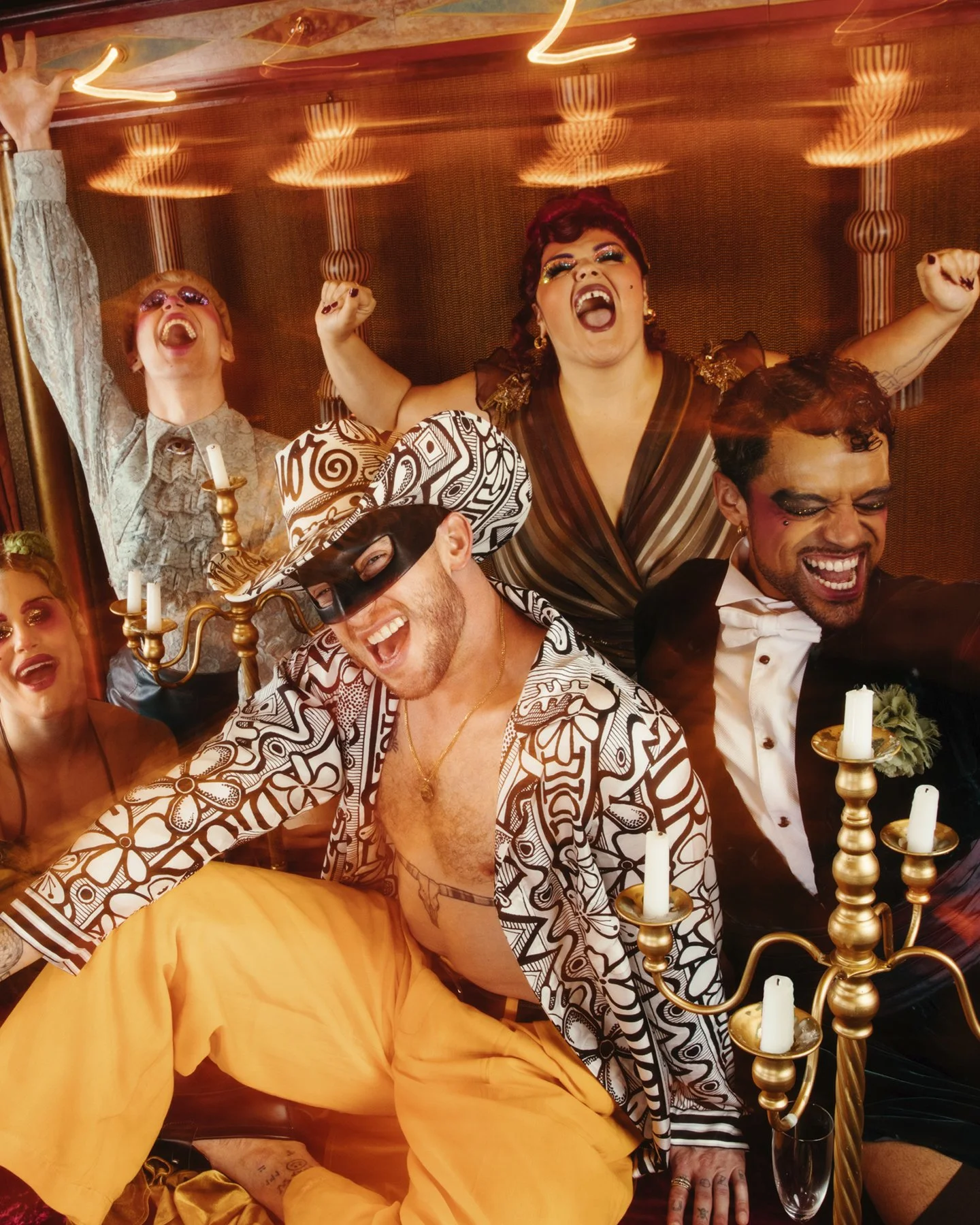 A group of friends celebrating at a party, dressed in colorful, expressive costumes, with several people smiling, cheering, and dancing. There are candles on candelabras and a lively, festive atmosphere.