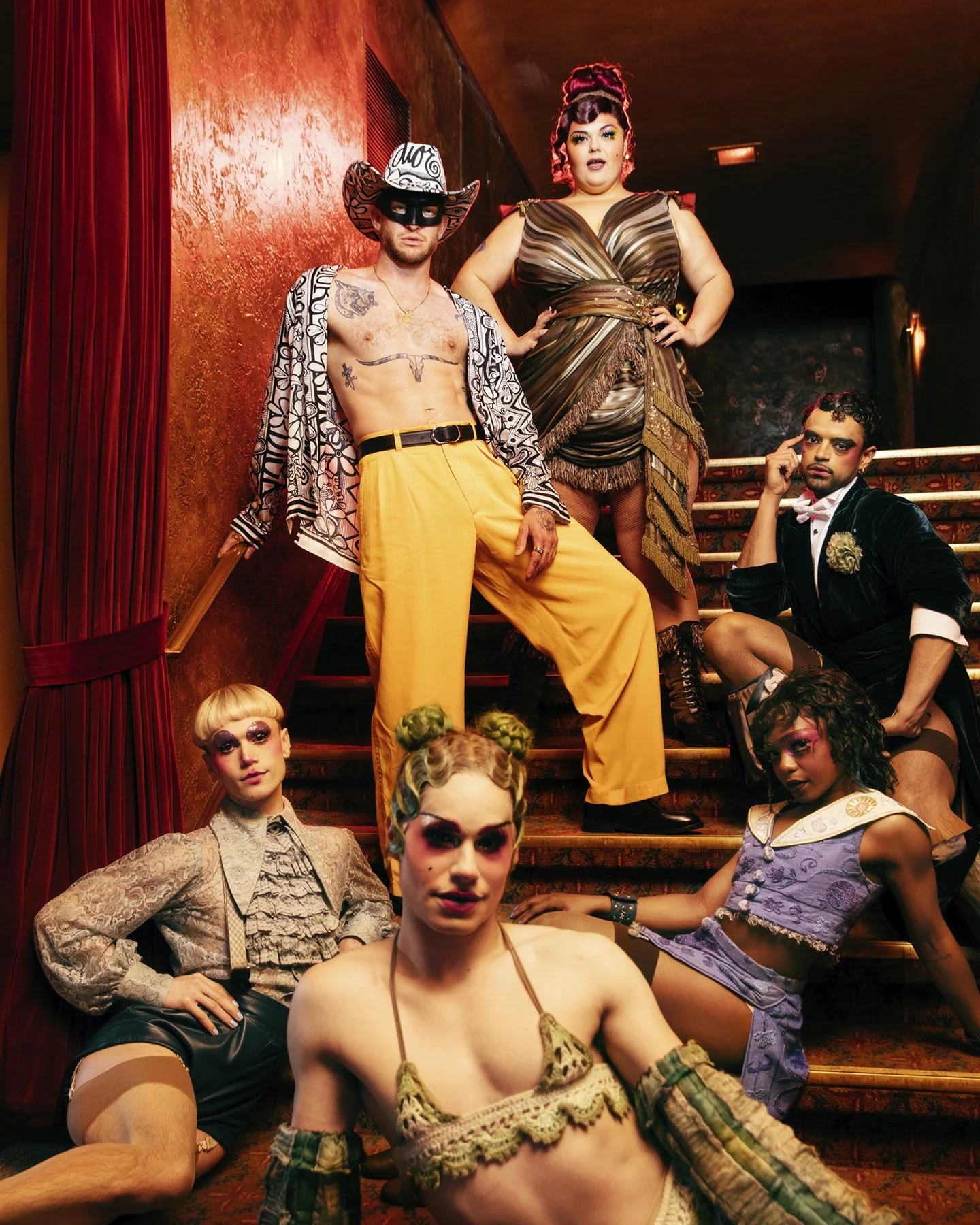 Group of diverse, flamboyantly dressed individuals posing on a staircase and floor in an ornate, dimly lit interior, with vibrant makeup and bold fashion styles.