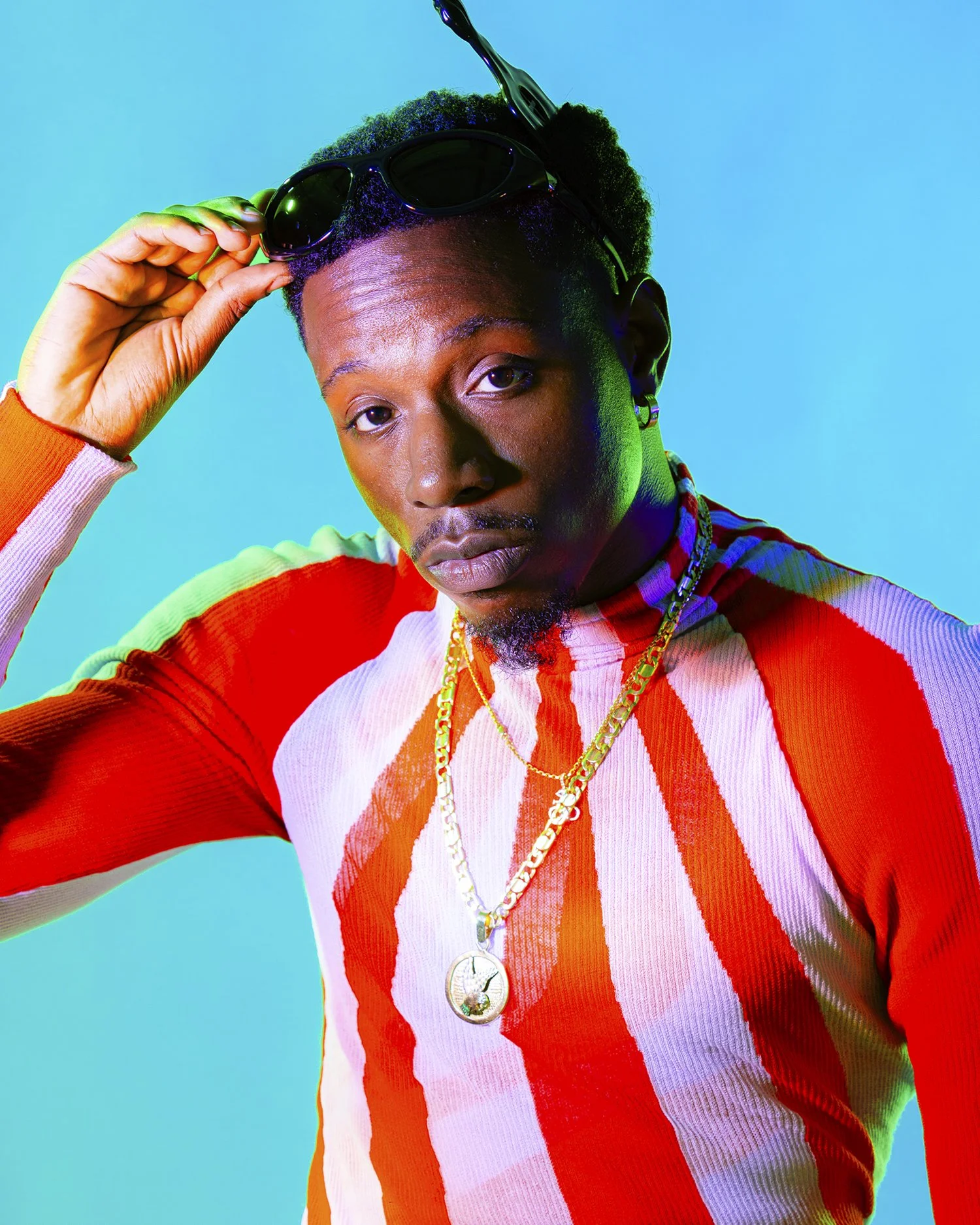 A man wearing a red and white striped long-sleeve shirt, gold chains, and black sunglasses on his head, posing against a blue background.