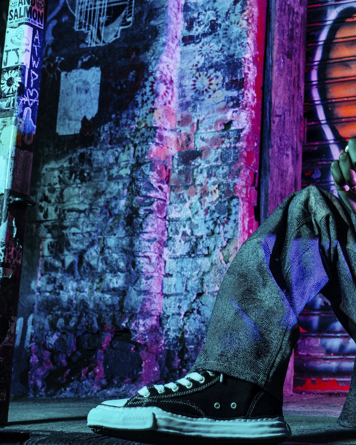 Close-up of a person's leg wearing a black and white sneaker, with jeans and a jacket, against a colorful, illuminated brick wall background.