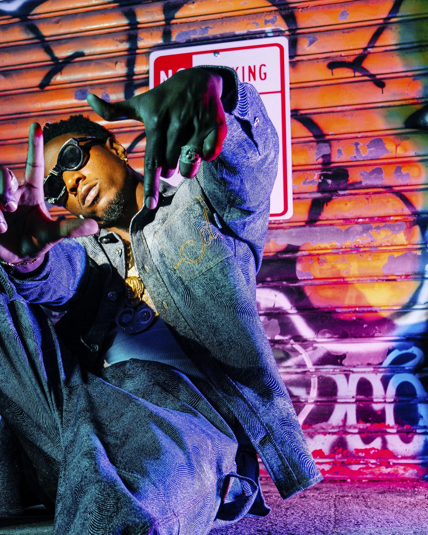 A person wearing sunglasses and a denim jacket making hand gestures in front of a colorful graffiti backdrop and a no parking sign.