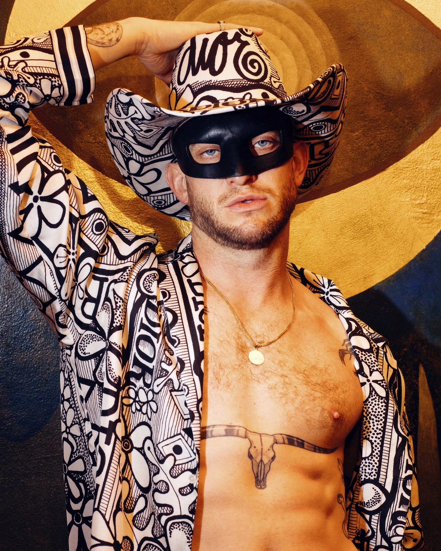 A man wearing a black mask, a large patterned hat, and an open shirt with a black and white abstract design. He is holding his hat with one hand, has a gold necklace, a tattoo of a bull skull on his chest, and a small tattoo on his shoulder. The back