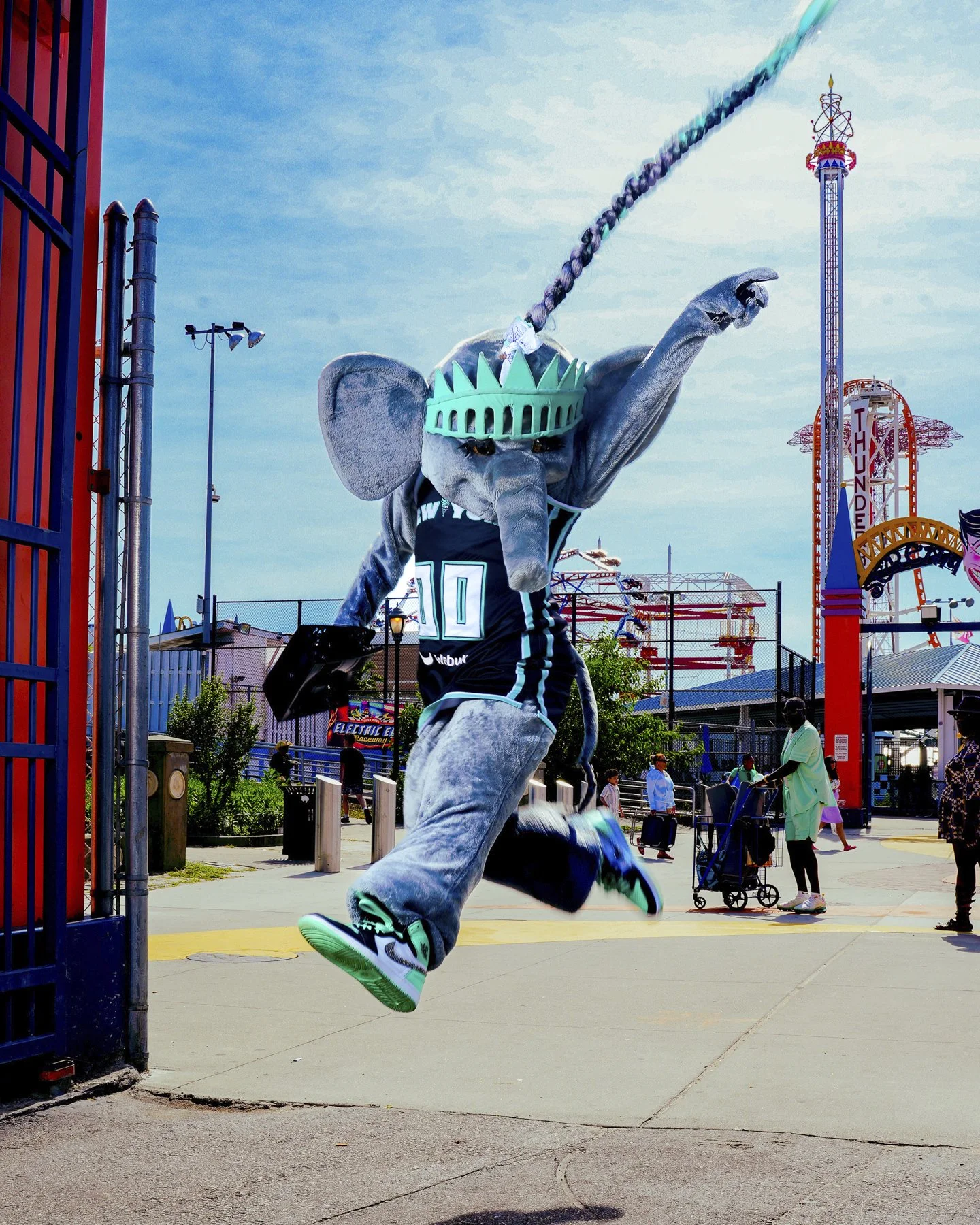 Person in an elephant mascot costume dressed in a sports jersey, jumping in front of an amusement park entrance with rides and people in the background.