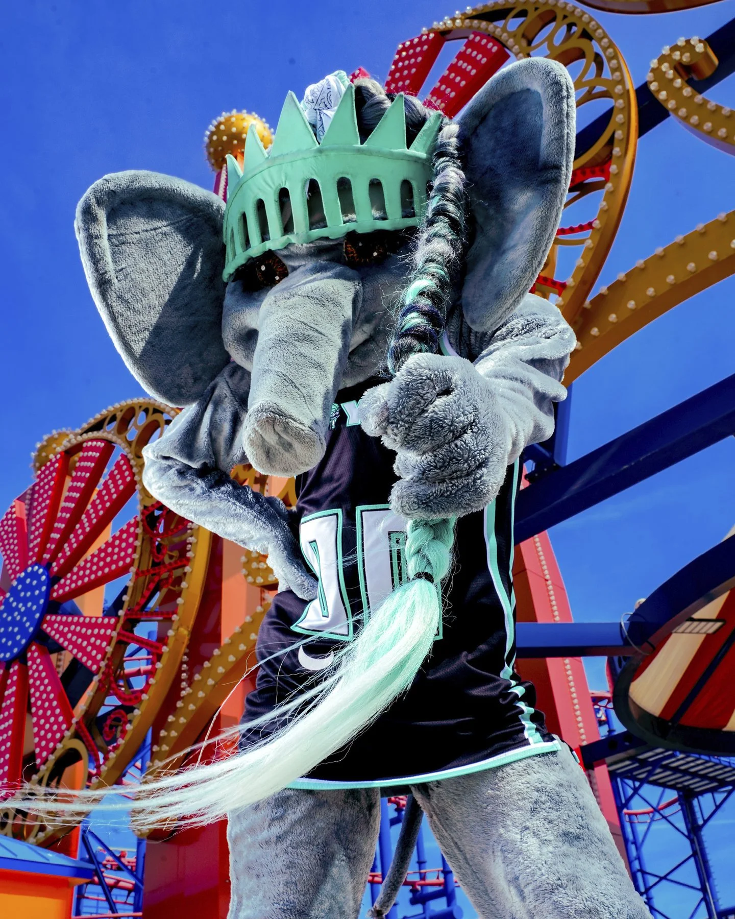 Person in a grey elephant costume with a green crown and braid, standing in front of a colorful amusement park ride with red, yellow, and blue parts, under a clear blue sky.