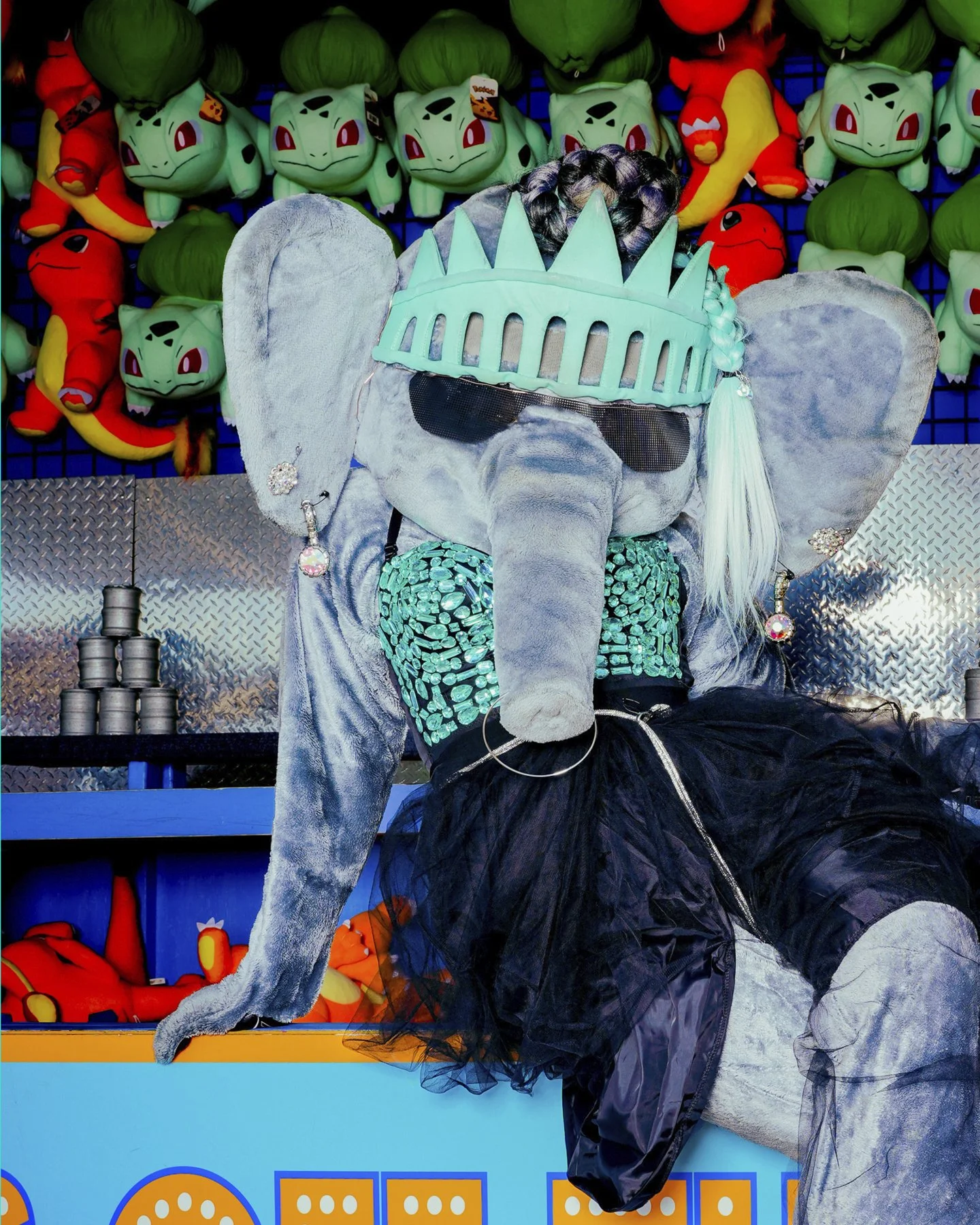 A person dressed in an elephant costume with sunglasses, a turquoise headband resembling the Statue of Liberty, and a black tulle skirt, sitting in front of a wall of Pokémon plush toys.