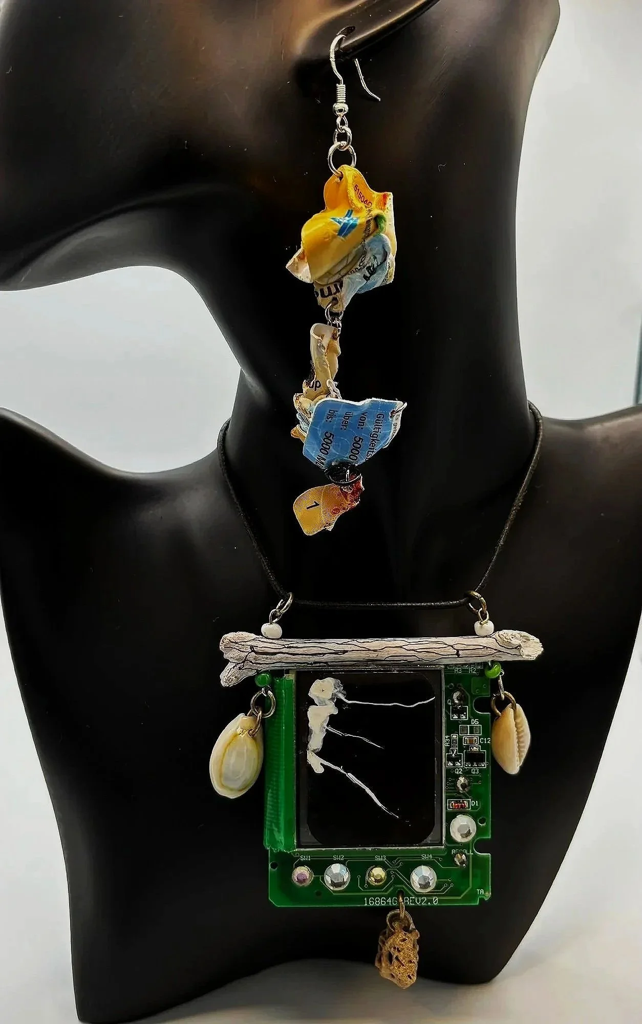 Halskette und Ohrring an einem schwarzen Modell. Die Halskette besteht aus elektronischem Bauteil, Muscheln, einem Holzstück und anderen kleinen Gegenständen. Der Ohrring enthält einen zerknüllten Geldschein.
