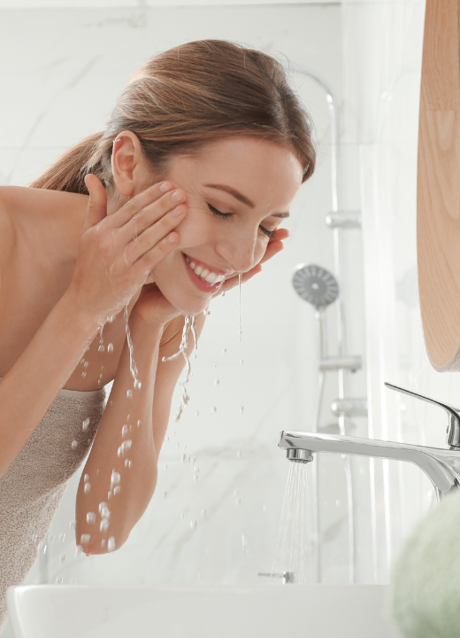 A woman rinses her face in a bathroom sink with water from a faucet.