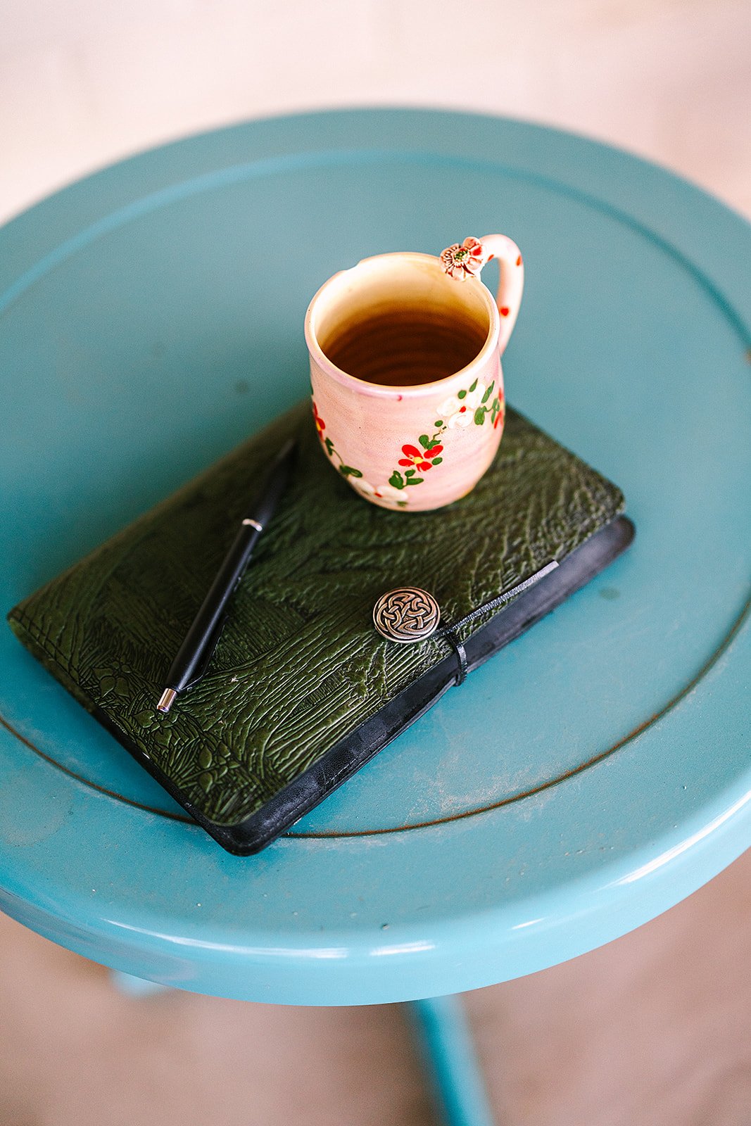 A top view of a pink floral mug filled with a hot beverage, placed on a textured green notebook, with a black pen on a blue round table.