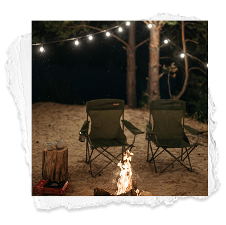 Two camping chairs around a campfire at night, with string lights overhead and trees in the background.