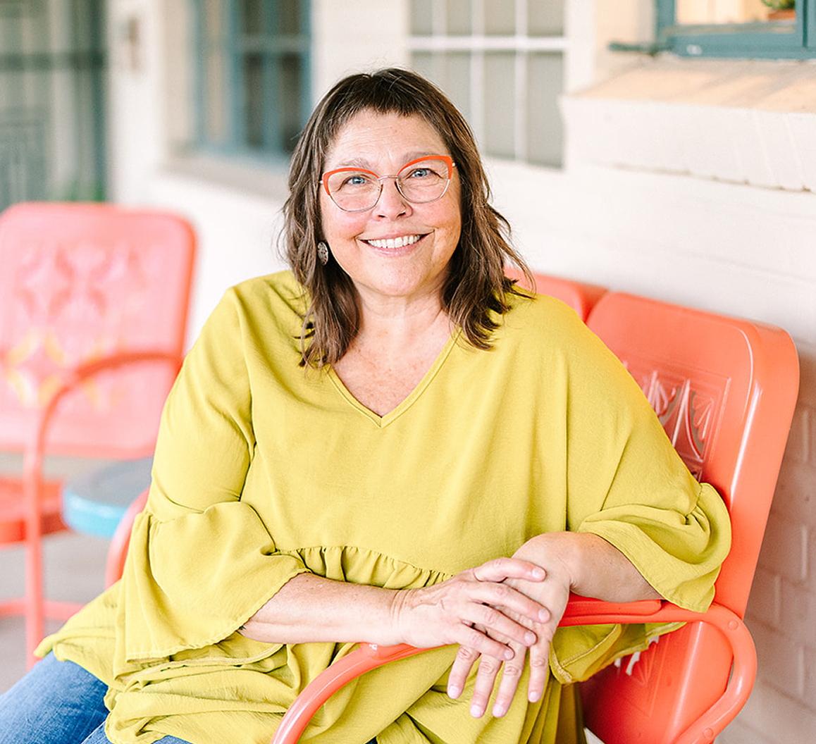 A smiling woman with glasses, wearing a yellow top, sitting on an orange chair in an outdoor setting with colorful furniture.