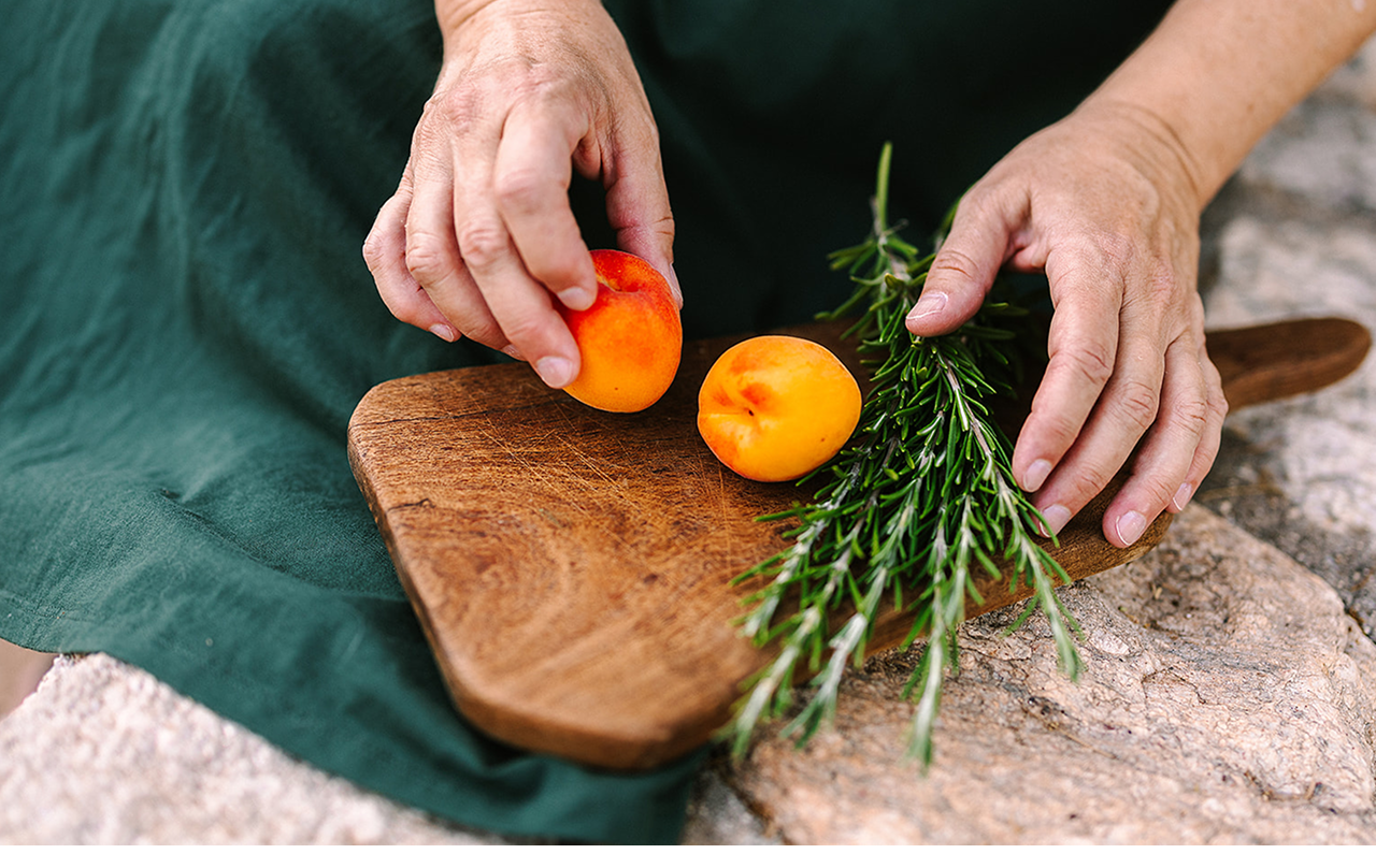 Hands arranging apricots and sprigs of rosemary on a wooden cutting board