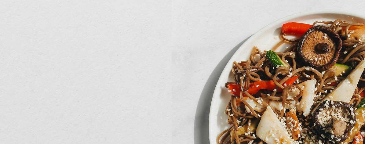 A plate of stir-fried noodles with sliced mushrooms, red bell peppers, zucchini, and sesame seeds.