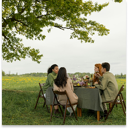 Four women sitting at a table outdoors beneath a large tree, enjoying a meal together in a grassy field.