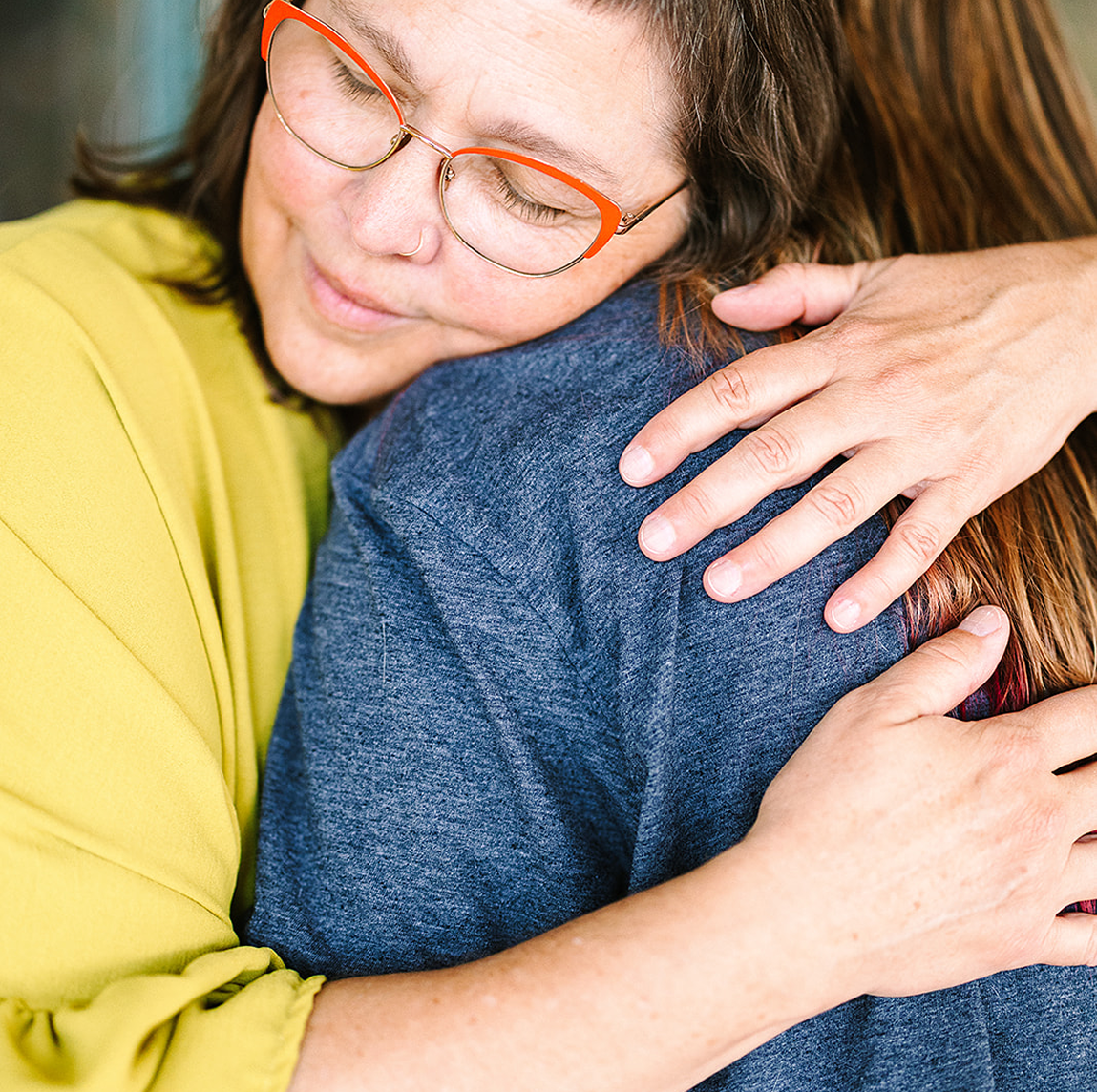 Woman with glasses hugging another person, both with their eyes closed, expressing comfort and warmth.