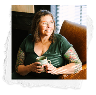 Smiling woman with glasses and tattoos holding a mug, sitting at a wooden table near window blinds.