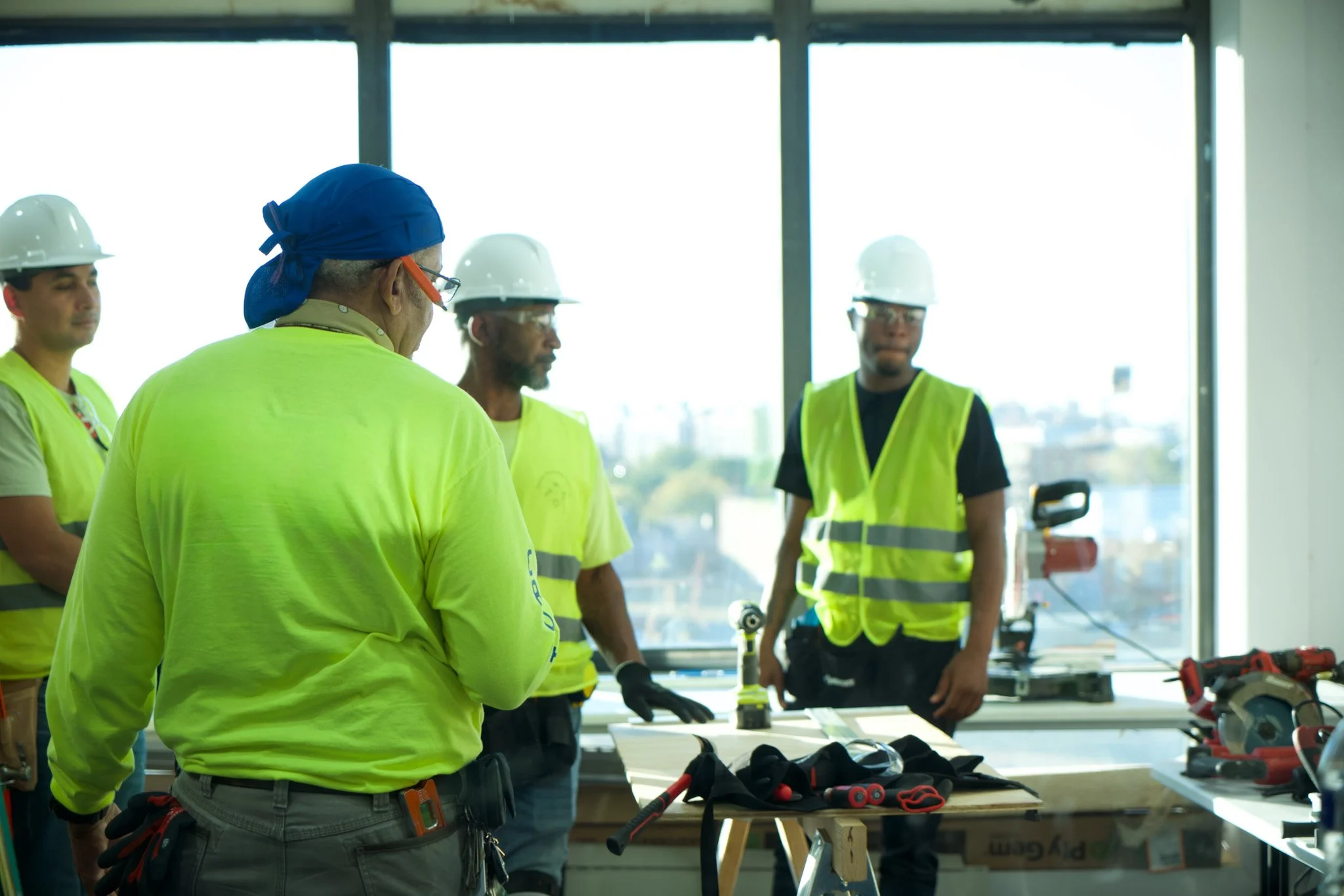Construction workers wearing safety vests and helmets in an indoor setting with large windows, gathered around a table with tools and equipment.