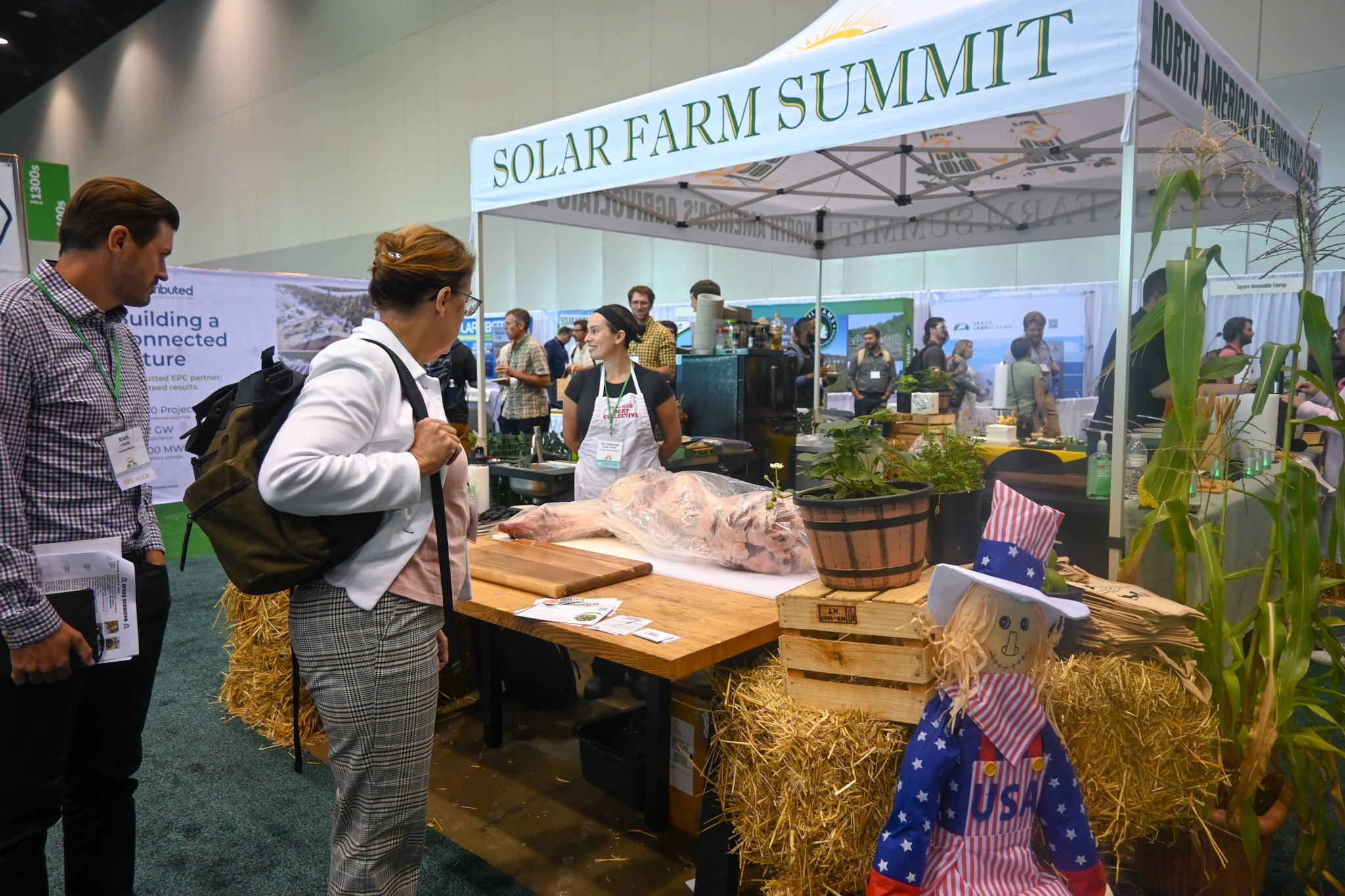 People visiting a booth at the Solar Farm Summit, with a display of a pig carcass, plants, and decorations, including a scarecrow dressed in patriotic American clothing.