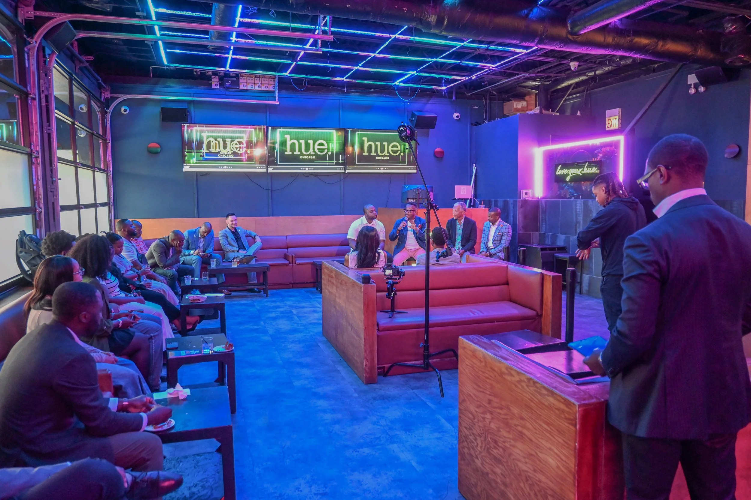 Inside a vibrant lounge or event space with neon lights, where a panel discussion or interview is taking place. Several people are seated on a long, curved sofa and some are standing, engaging with microphones. Attendees are seated along the walls, watching the event, with some taking notes or pictures. The decor features purple and blue neon lighting, with signs that say 'hue' and a neon frame that reads 'loving hue' on the wall.