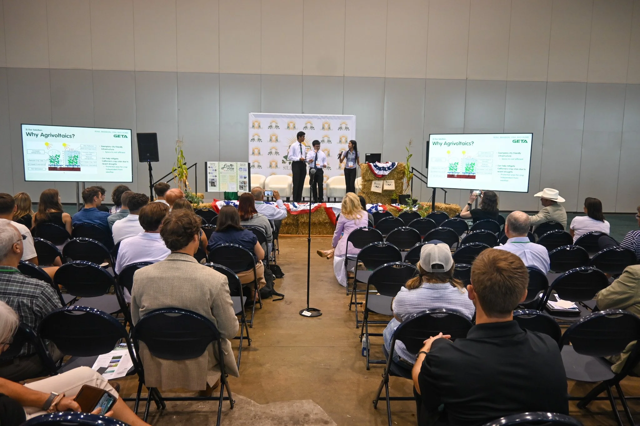 A conference or seminar with an audience seated facing a stage where three people are presenting. The stage has two large screens displaying slides titled 'Why Agrivoltaics?' and decorated with patriotic banners and hay bales.