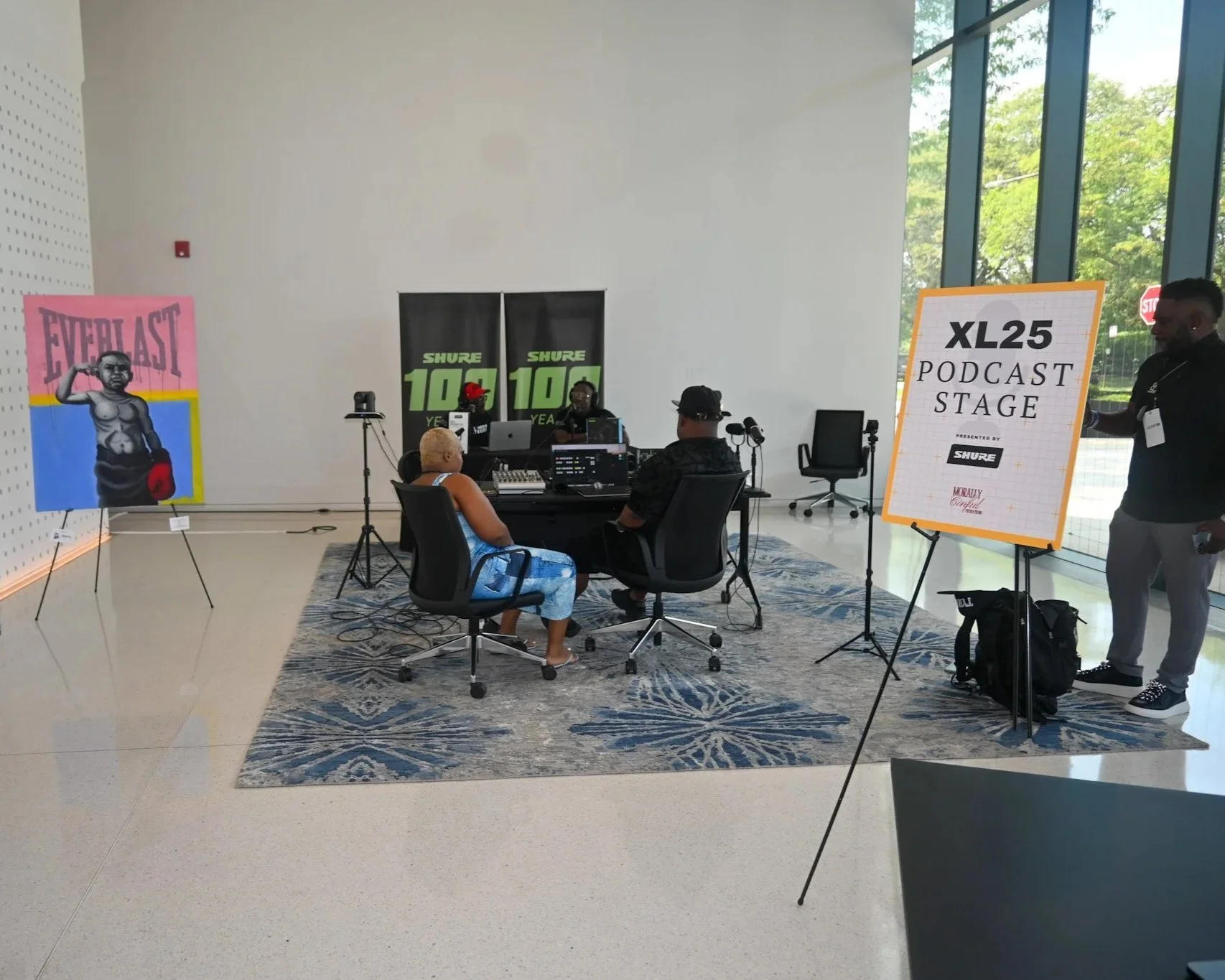 Podcast recording studio with two hosts seated at a table, surrounded by equipment, and a woman in blue sitting nearby. There is a large sign on an easel that reads XL25 Podcast Stage, with a smaller sign promoting Shure.