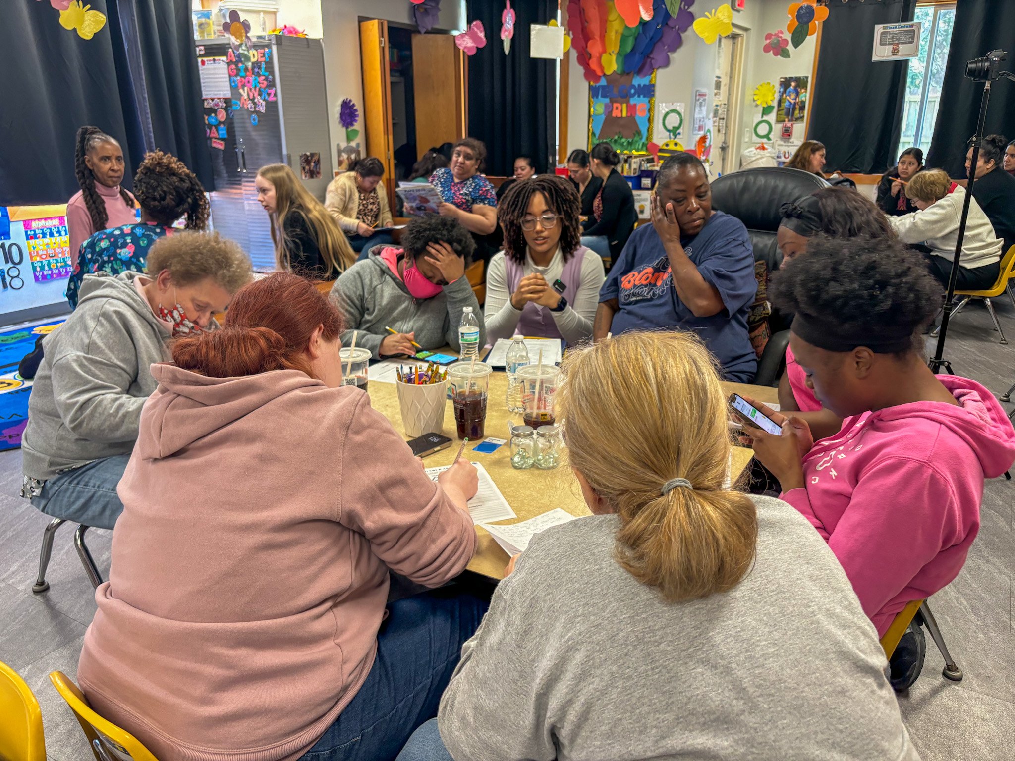 A group of women gathered around a table indoors, engaged in conversation, with some looking at phones and notes. The background features colorful decorations, including flowers and banners, with additional people seated at tables.