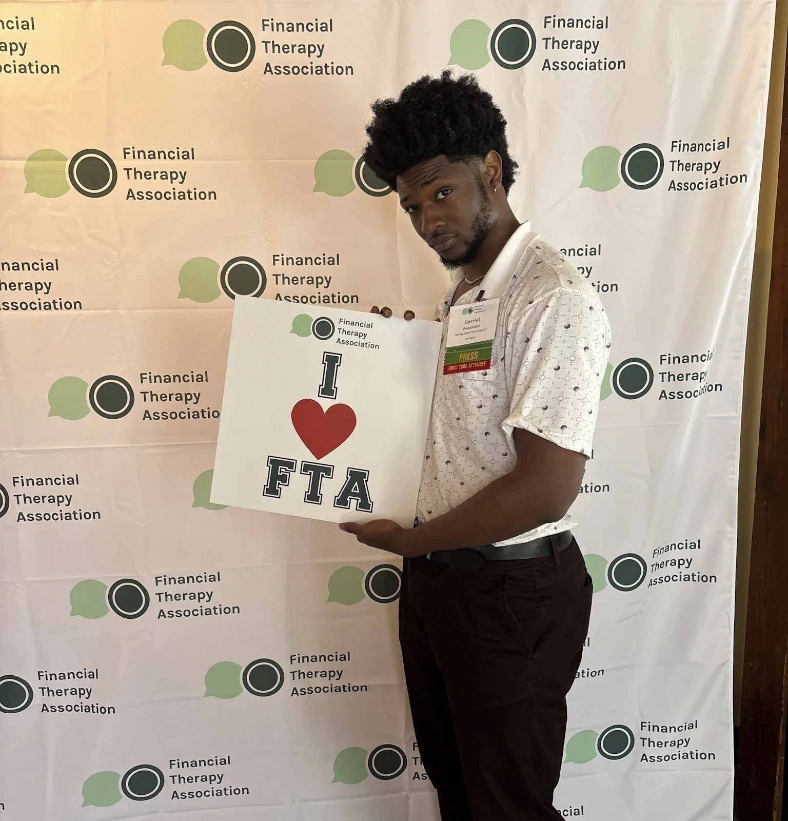 A young man with curly hair holds a white sign that says "I love FTA" with a red heart symbol. He is standing in front of a backdrop with a repeating logo and text for the Financial Therapy Association. The man is wearing a white short-sleeve button-up shirt with small black dots and black pants, and has a name badge with a green "Press" tag.
