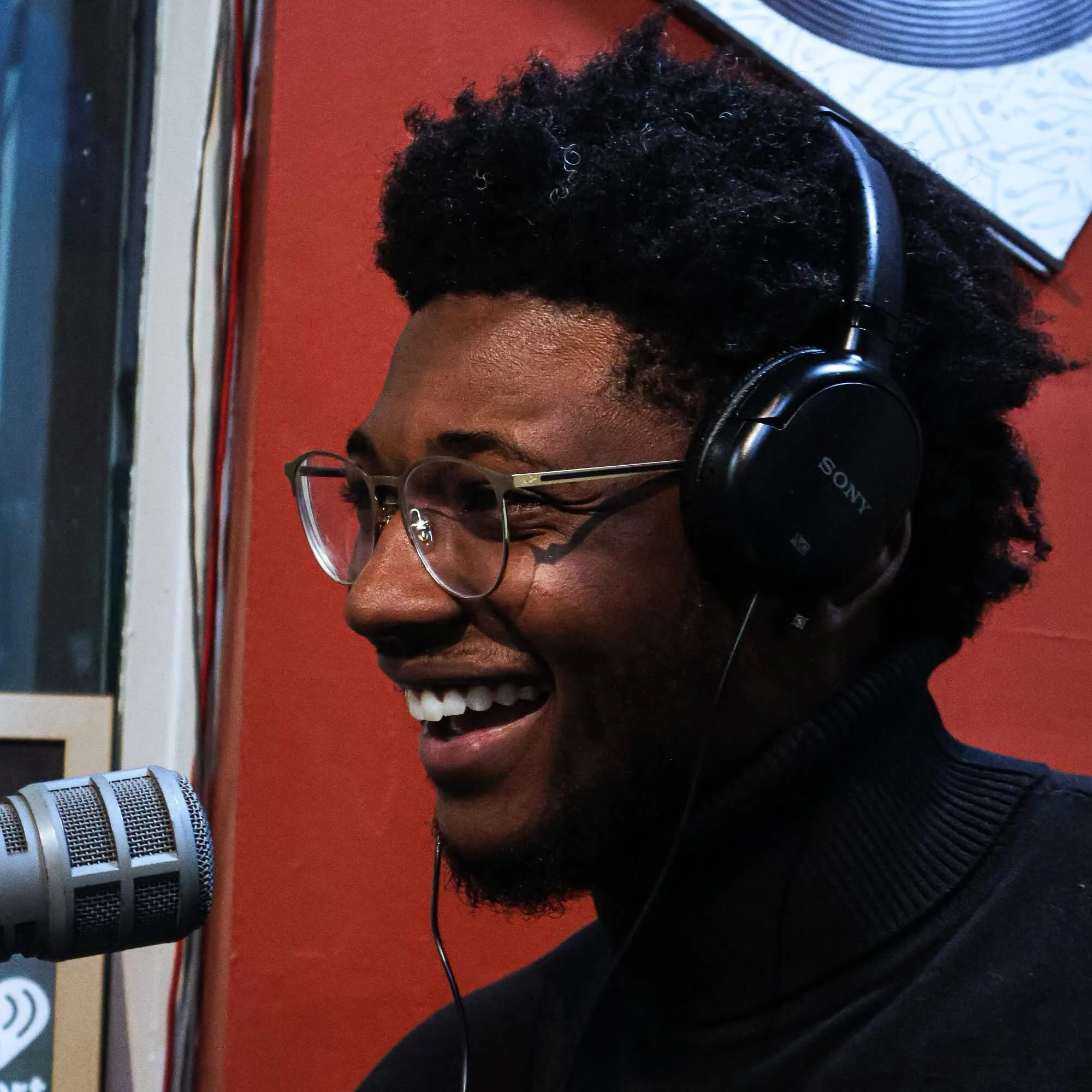 A young man with glasses and curly black hair wearing headphones, smiling at a microphone in a recording studio.
