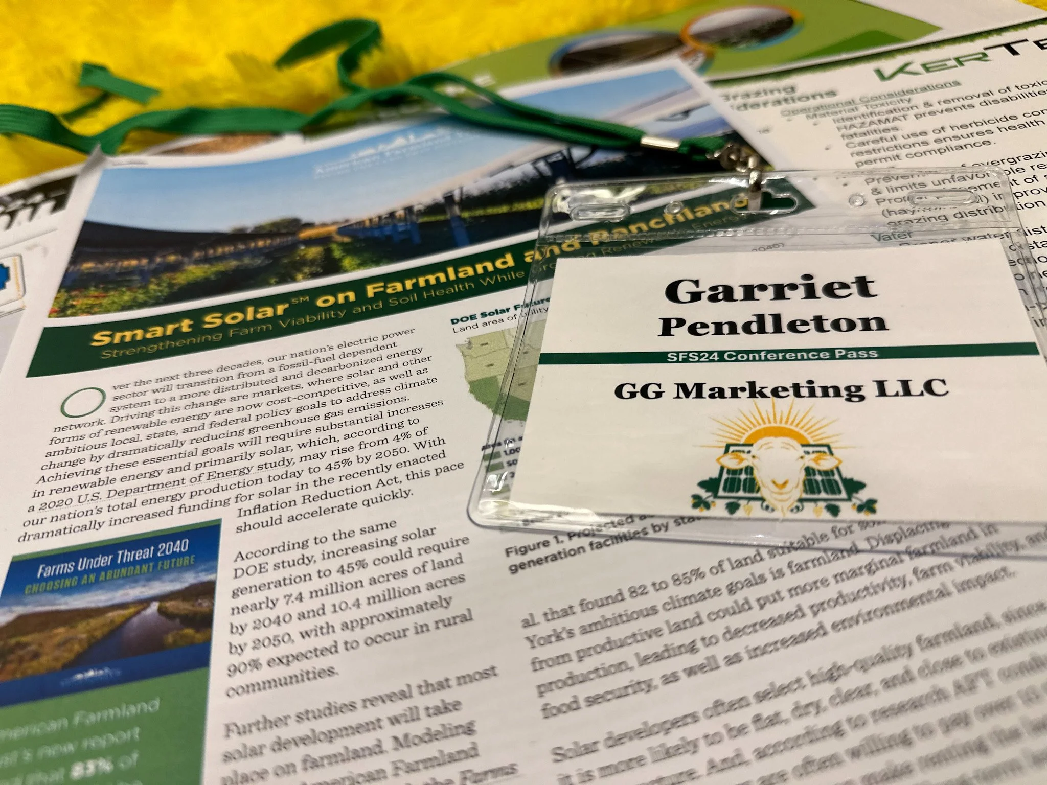 A conference pass for Garriet Pendleton from GG Marketing LLC placed on a document, with a solar-themed logo featuring a sheep and solar panels, alongside brochures about farmland and solar energy.