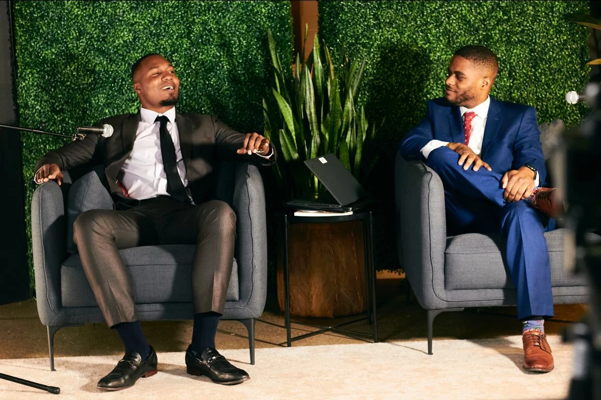 Two men in suits sitting on gray armchairs having a conversation with a green leafy wall behind them.