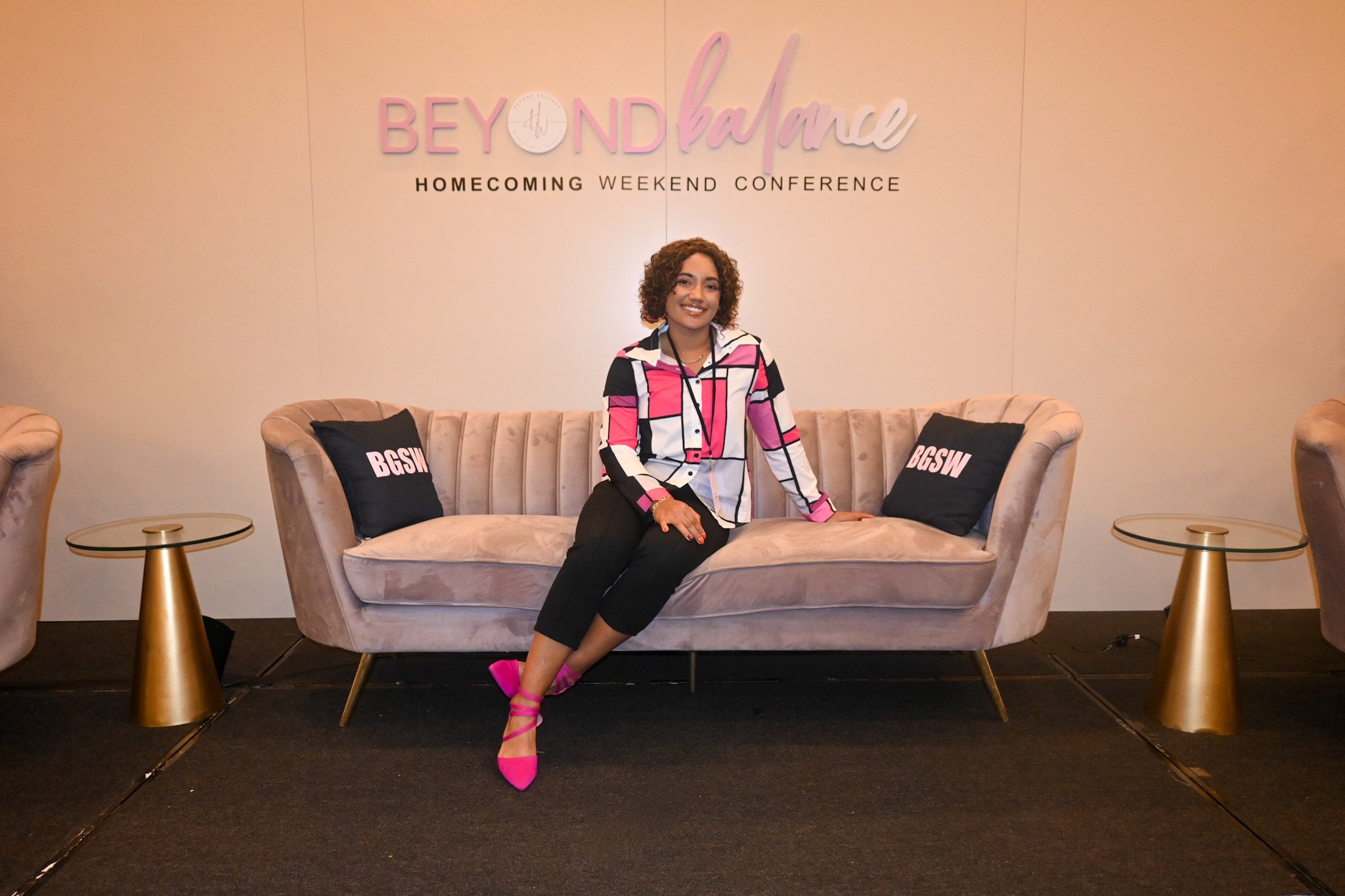 A woman with curly hair sitting on a beige velvet sofa in front of a wall with the text 'BEYOND balance' and 'HOMECOming WEEKEND CONFERENCE.' She is smiling, wearing a pink and white checkered shirt with black pants and pink heels. There are black pillows with white text 'BGSW' on each side of her.
