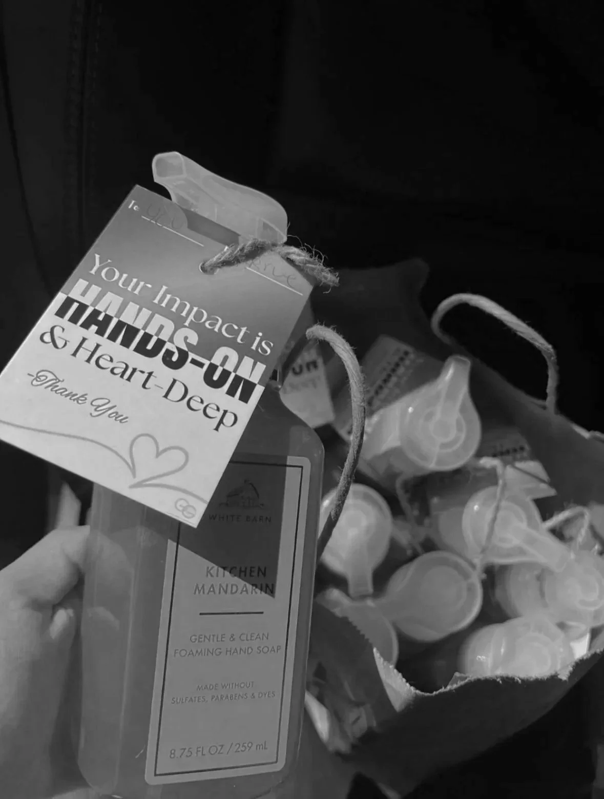 A hand holding a bottle of Wooden Barn kitchen mandarin foaming hand soap, with a thank you card attached that says 'Your impact is HAM-S-ON & Heart Deep' and a heart illustration. In the background, there's a paper bag filled with numerous pump bottles of soap.
