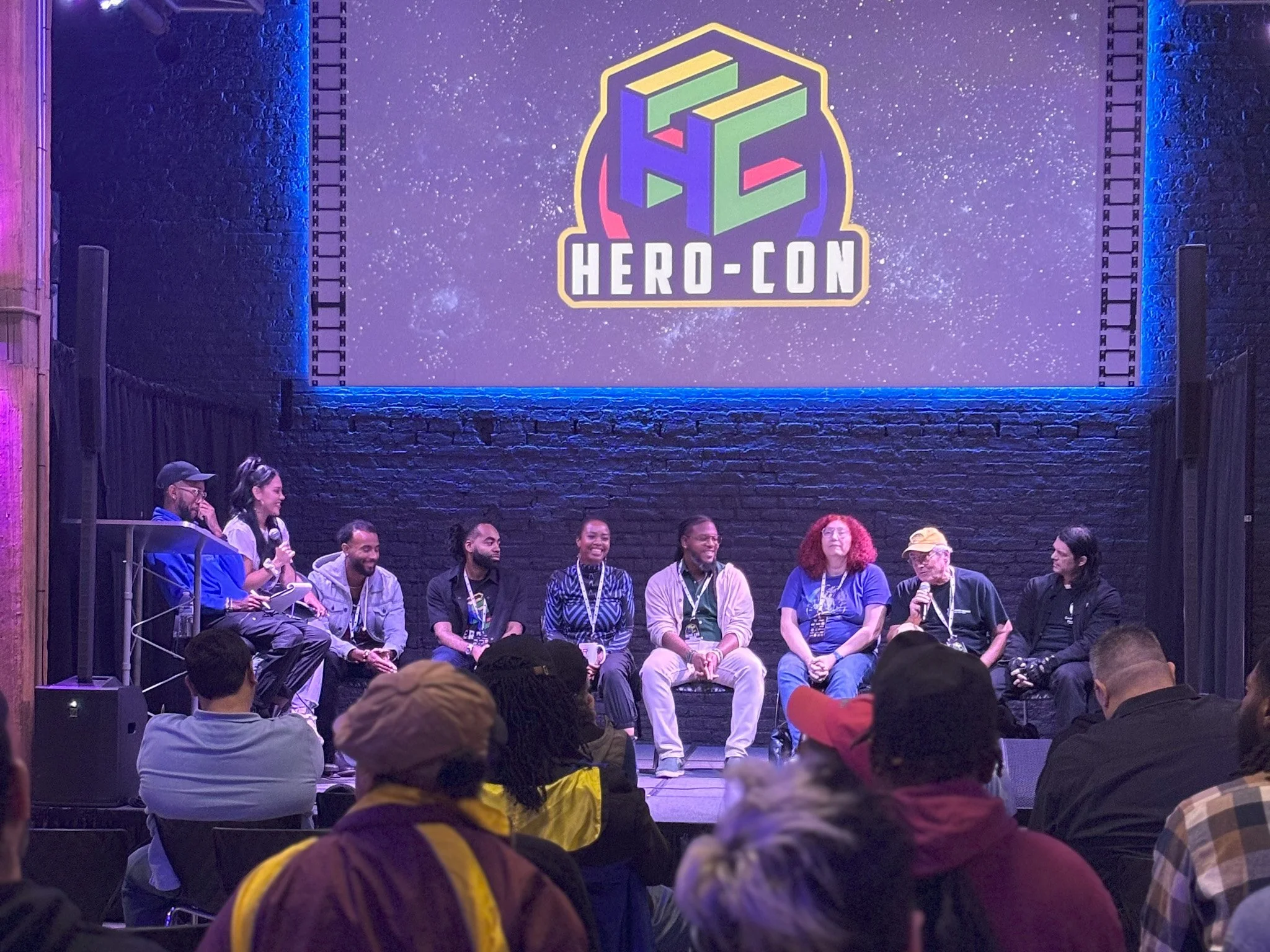 Panel discussion at Hero-Con, with nine people seated on stage under a large screen displaying the Hero-Con logo against a starry background. The audience is watching.