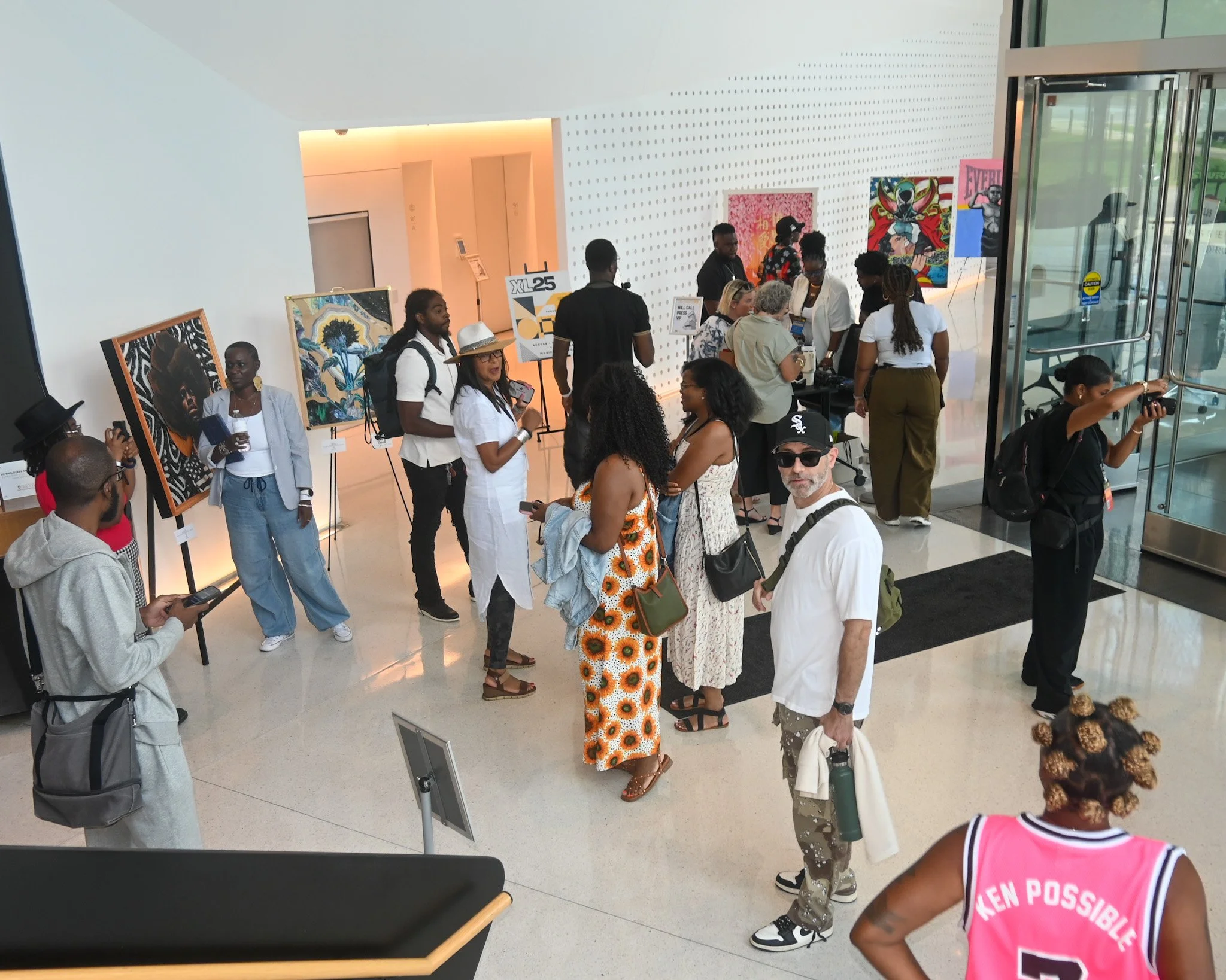 People attending an art gallery exhibition, viewing paintings on display, with some taking photos and others engaging in conversation, in a modern, well-lit space.