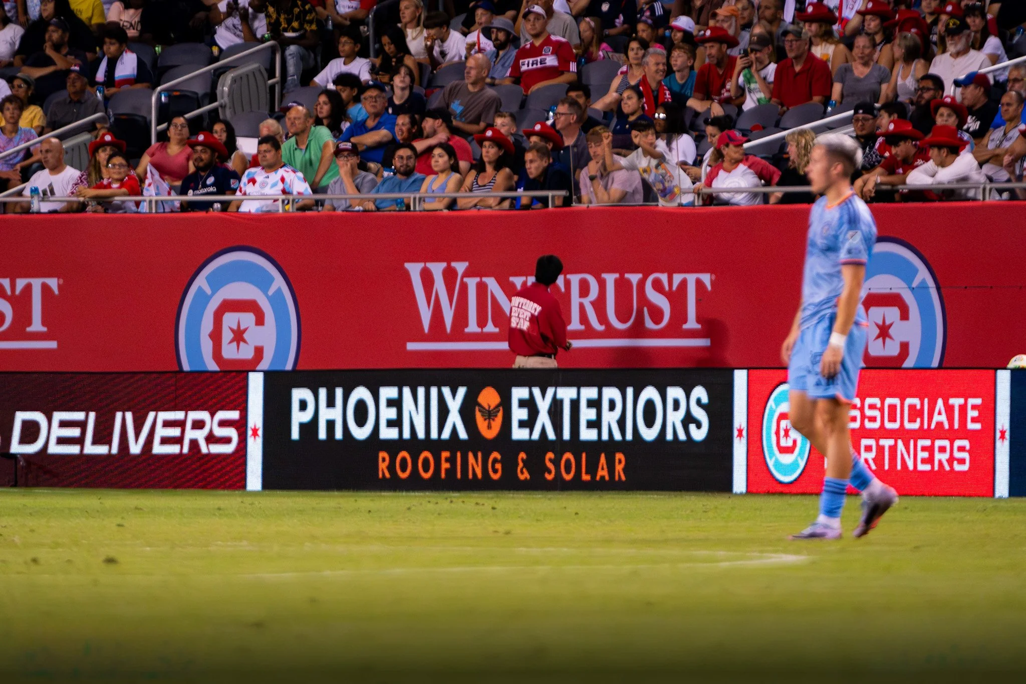 A soccer player in a light blue uniform stands on the field during a game, with a large crowd of spectators in the stands behind a red advertising banner for Phoenix Exteriors Roofing & Solar. The crowd is diverse, with some wearing hats and team jerseys.