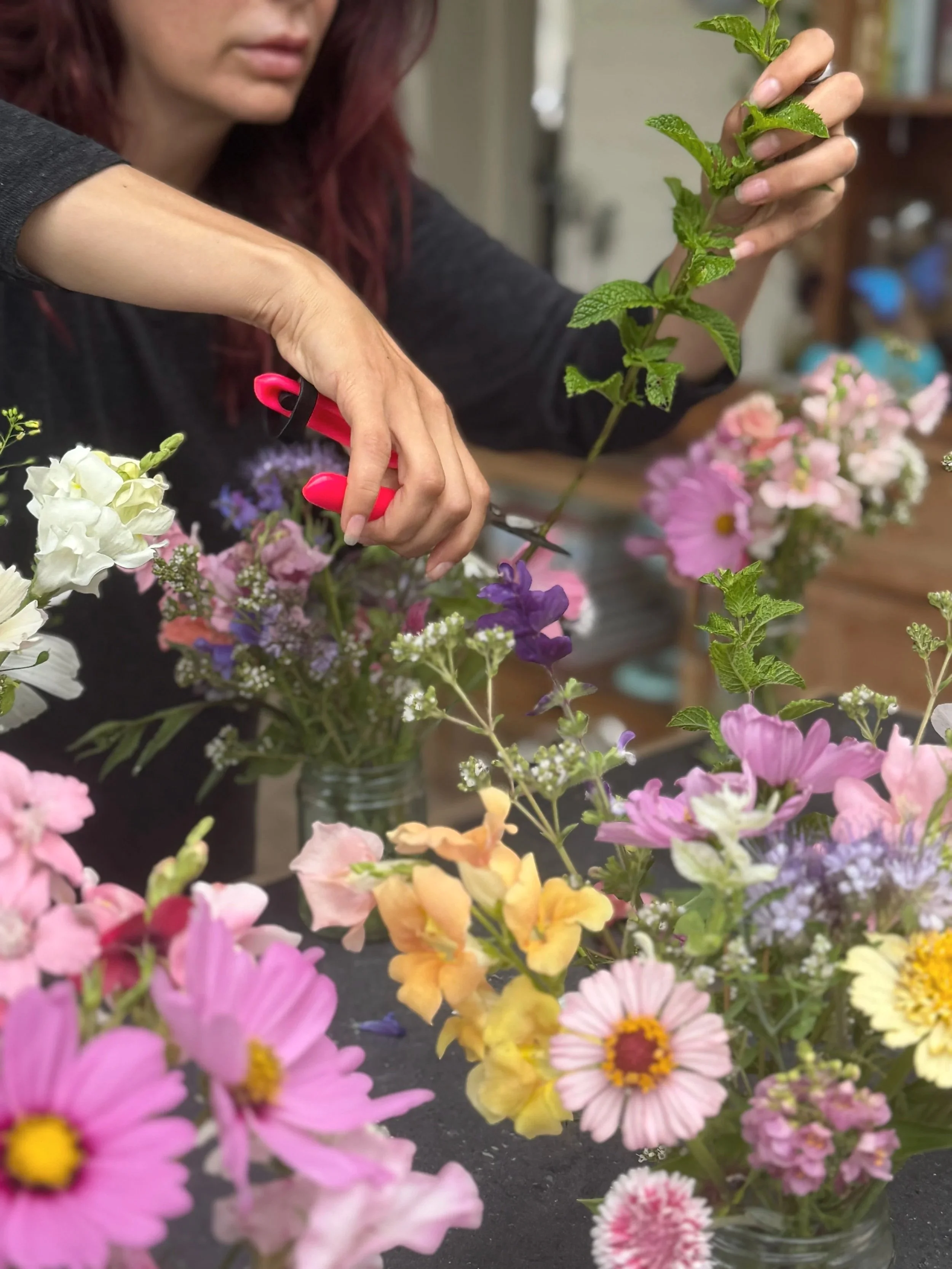 A person with red hair holds a pair of pink gardening scissors and arranges a bouquet of colorful flowers, including pink, purple, white, and yellow blossoms, on a dark surface with shelves in the background.