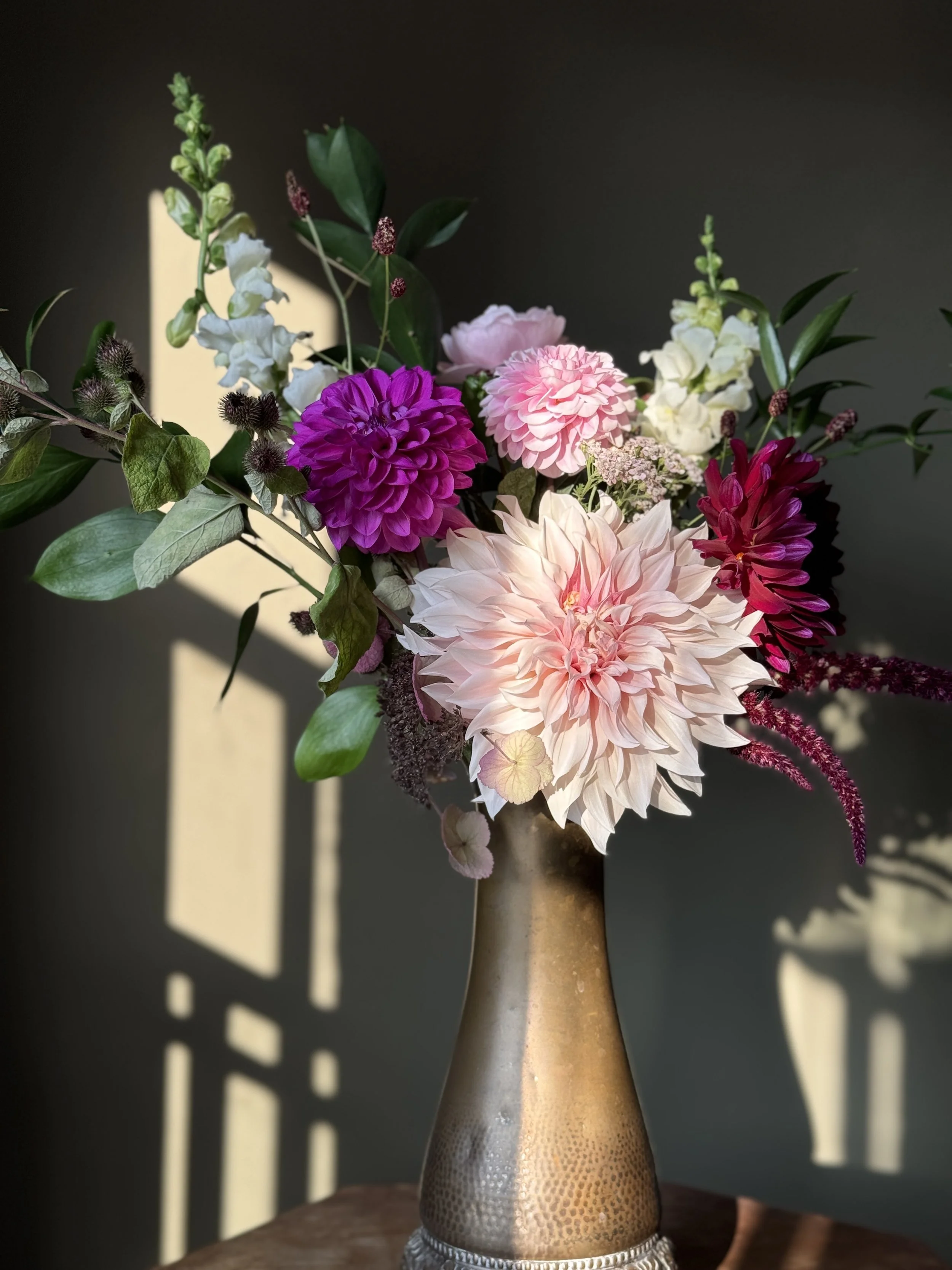 A flower arrangement in a metallic vase with a hammered pattern, featuring pink, purple, white dahlias, white roses, greenery, and other flowers, with shadows cast on a dark background.